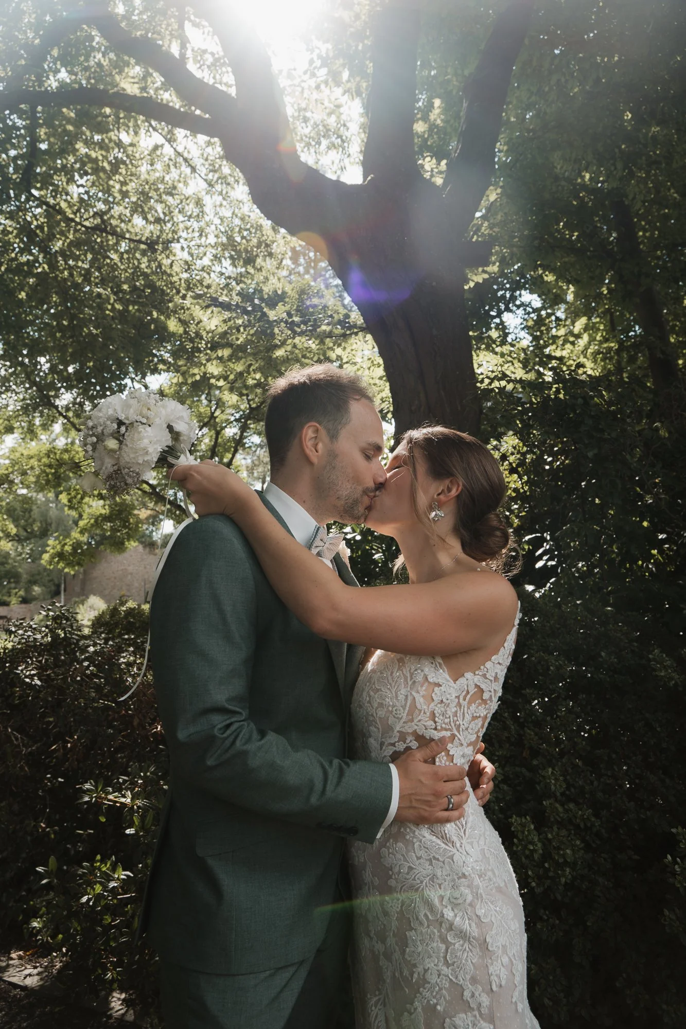 Ein Brautpaar küsst sich im Freien unter einem Baum, die Sonne scheint hindurch, die Braut trägt ein Spitzenkleid und der Bräutigam einen Anzug.