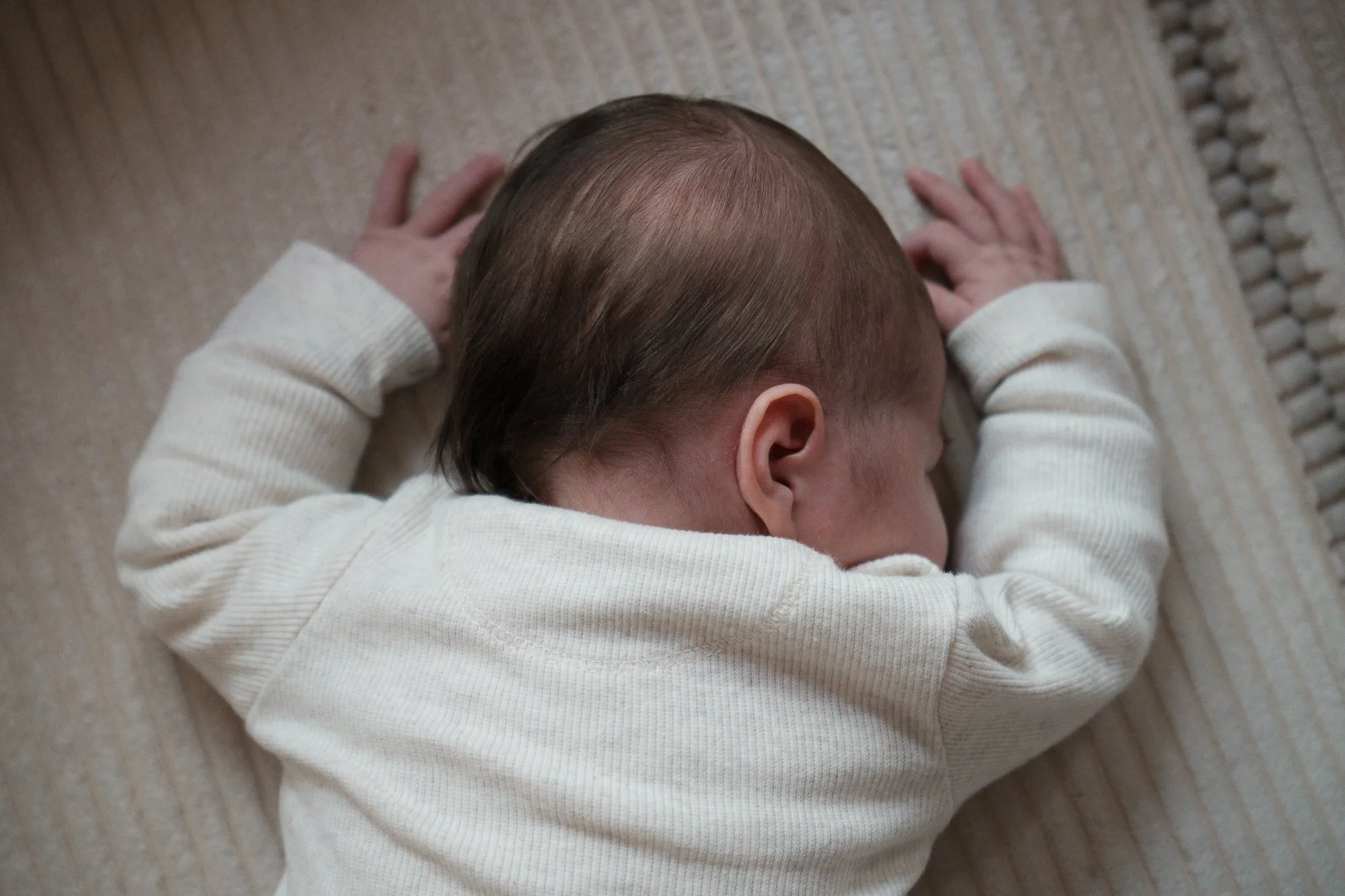 Ein kleiner Baby liegt auf dem Bauch auf einem Teppichboden, mit dem Kopf nach rechts, trägt ein weißes langärmeliges Kleid.