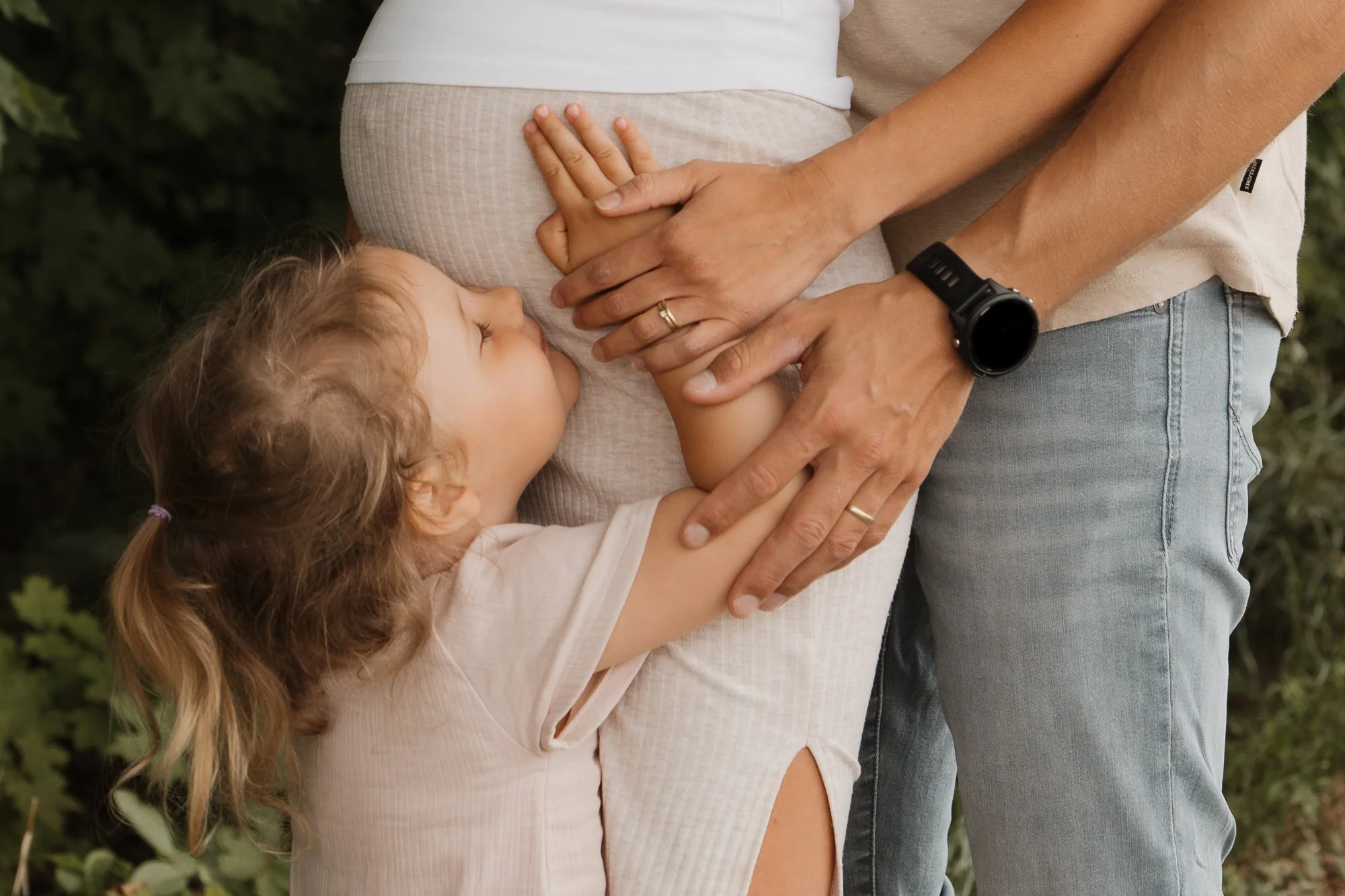 Ein kleines Mädchen klebt an einer schwangeren Bauch, während eine erwachsene Person sie liebevoll hält, im Freien, umgeben von Grün