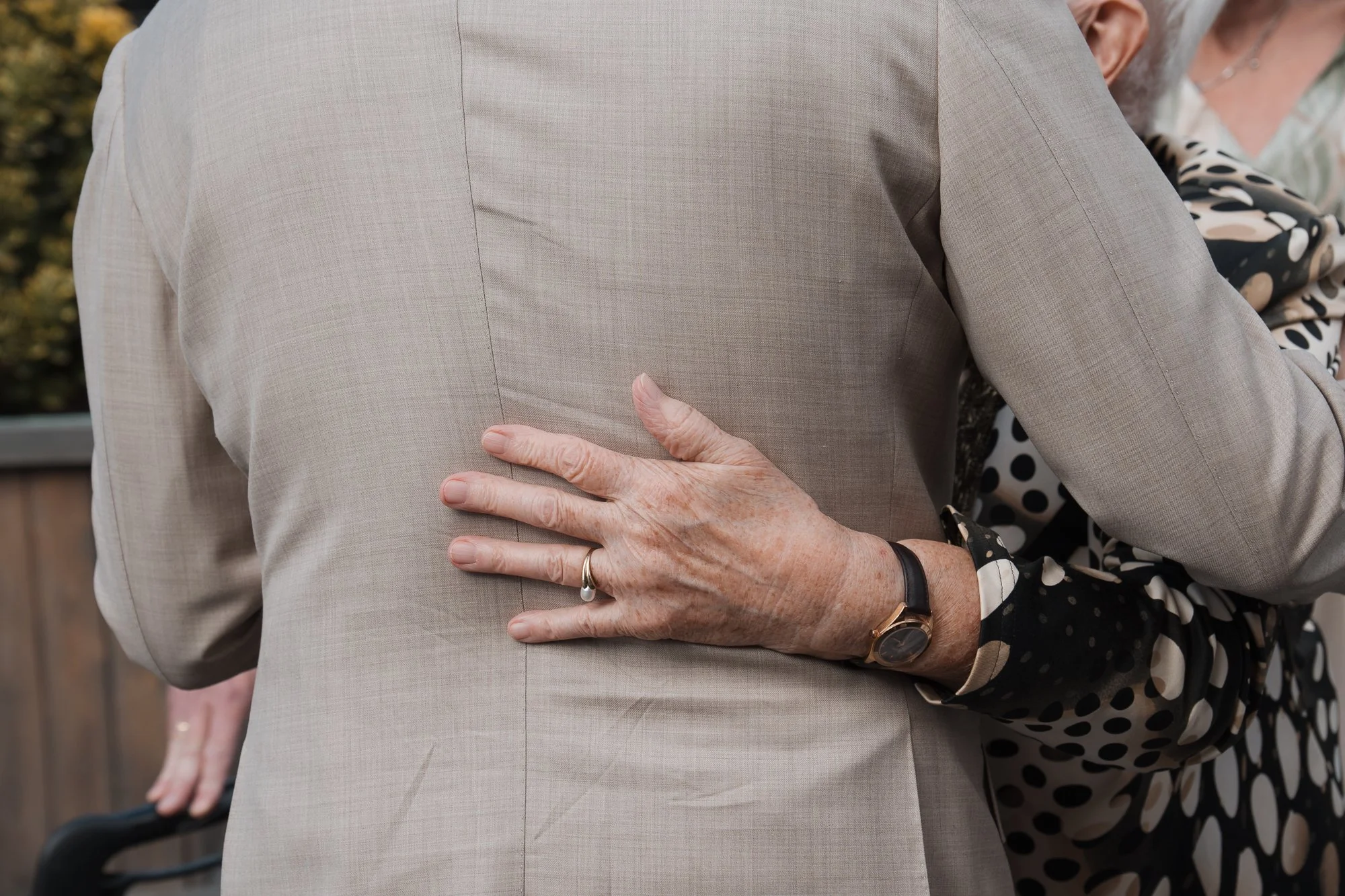 Eine ältere Frau hält die Hand an den Rücken eines Mannes, der einen grauen Anzug trägt, im Hintergrund eine weitere Person mit einem schwarzen Kleid mit weißen Punkten.