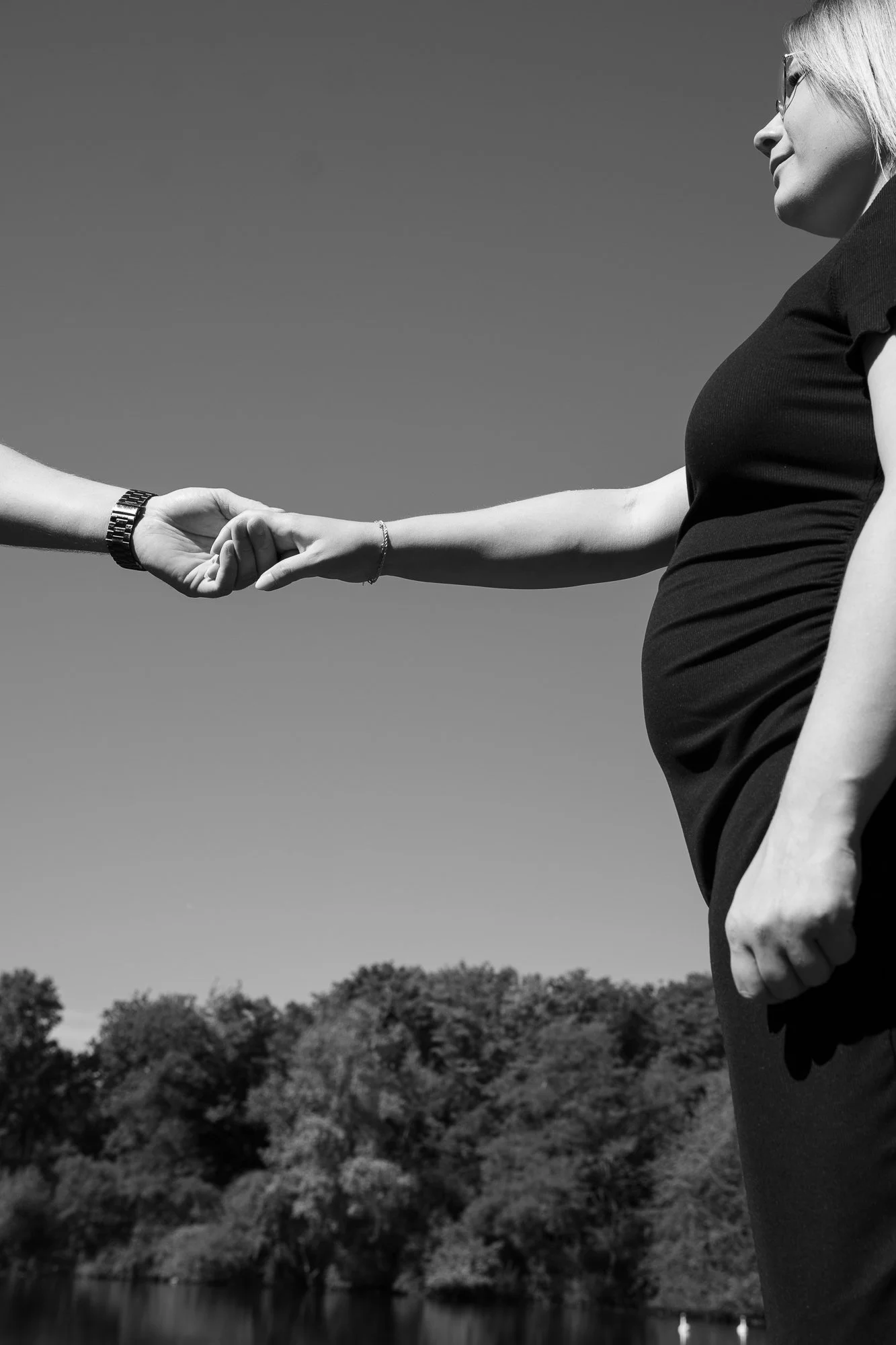 Schwangere Frau und Person reichen sich die Hand im Freien, Himmel und Bäume im Hintergrund, Schwarz-Weiß-Fotografie.