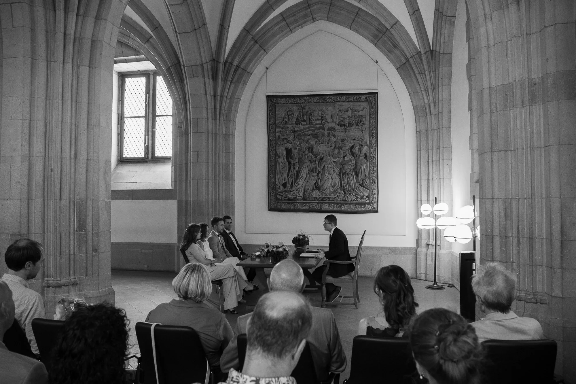 Eine Trauzeremonie in einer gotischen Kirche, mit Brautpaar, Trauzeugen, Ringträgern und einem Redner vor einem Publikum, im Schwarz-Weiß-Foto.