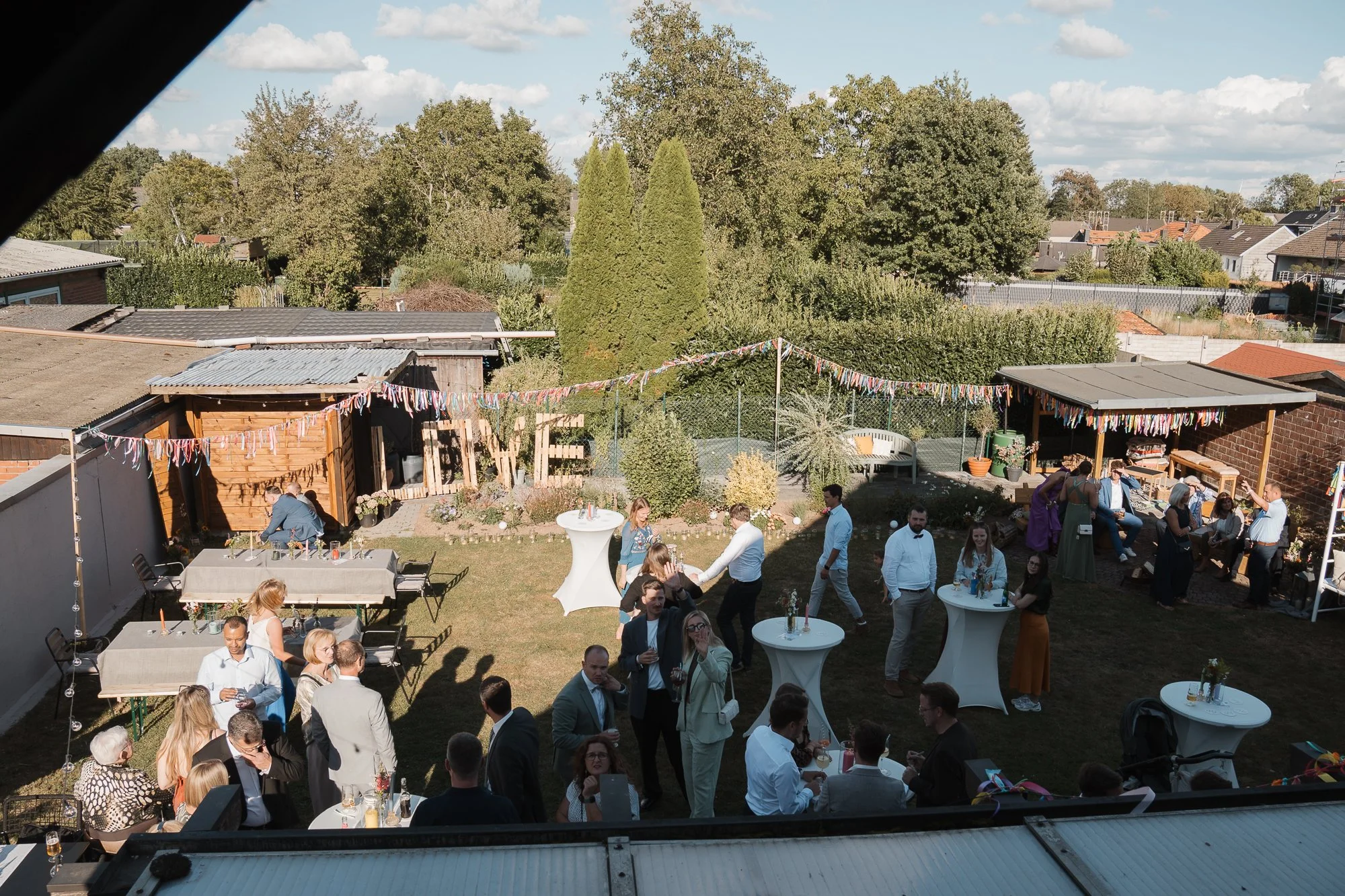 Menschen bei einer Gartenparty im Freien, bunt geschmückte Wäscheleine, Tische mit Getränken, Bauten und Bäume im Hintergrund