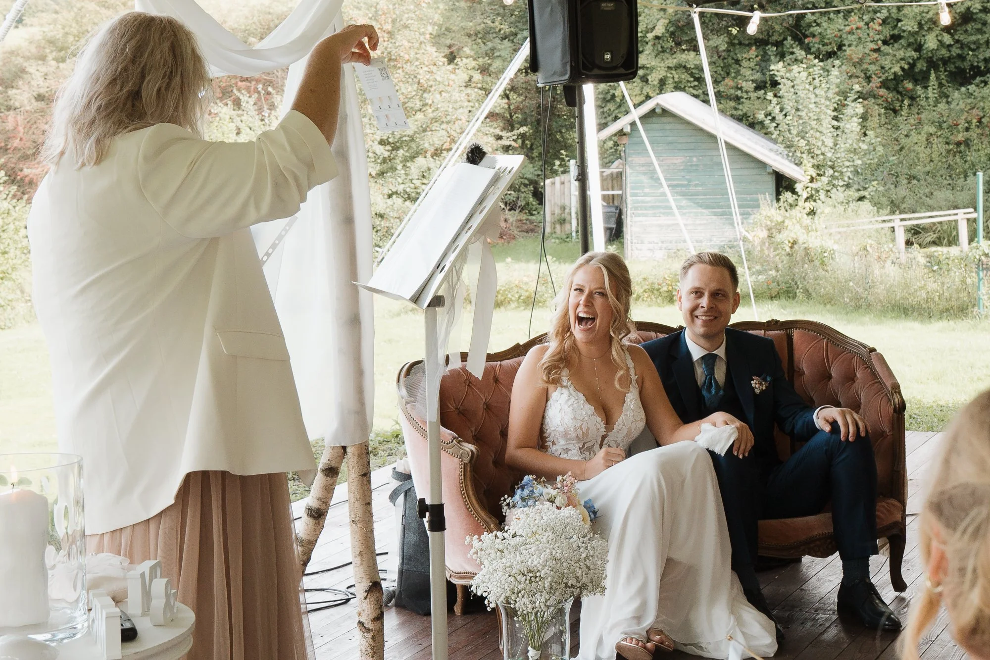Hochzeitspaar sitzt auf einer Couch im Freien während der Zeremonie, lachend und glücklich, mit Braut in weißem Kleid und Bräutigam in dunklem Anzug. Eine Person steht neben der Couch, hält einen weißen Stoff, vermutlich für die Zeremonie, und dekora