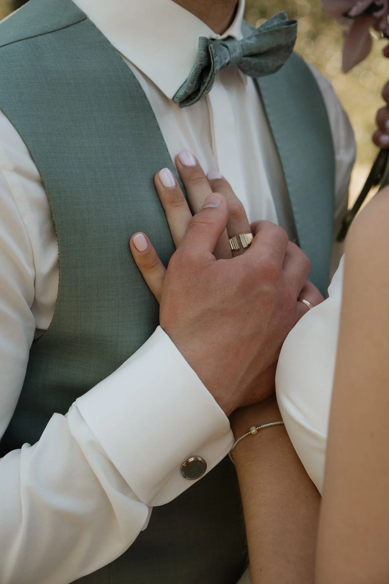 Nahaufnahme von einem Paar, das sich bei einer Hochzeit bei den Händen hält. Der Mann trägt einen grauen Anzug mit weißem Hemd und Fliege, die Frau trägt ein weißes Kleid und eine Perlenarmband. Beide tragen Eheringe.