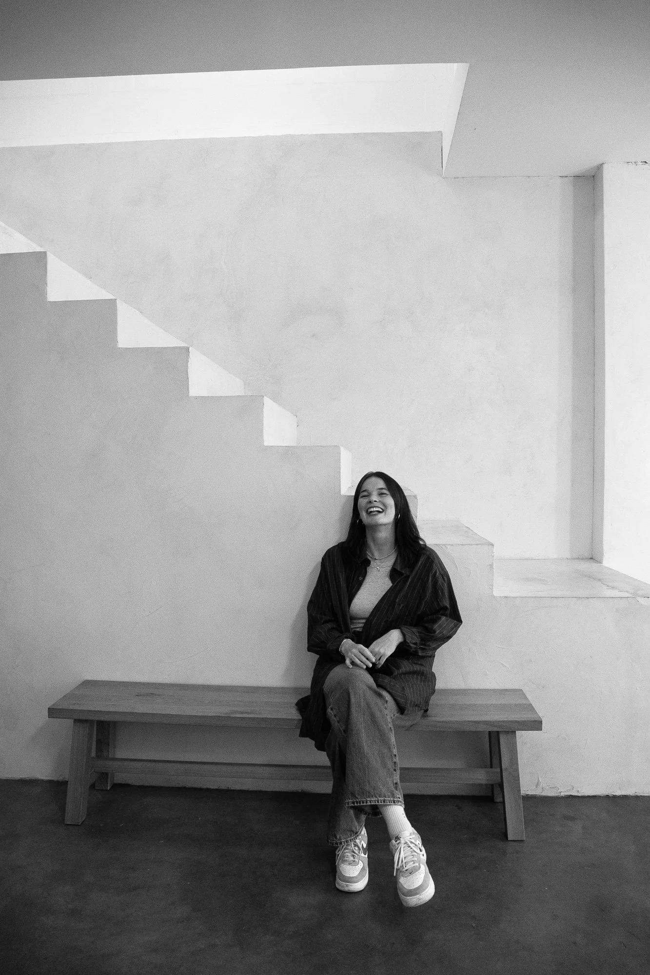 Eine lachende Frau sitzend auf einer Holzbank in einem minimalistischen Raum mit weißen Wänden und Treppenansicht im Hintergrund. Schwarz-Weiß-Fotografie.