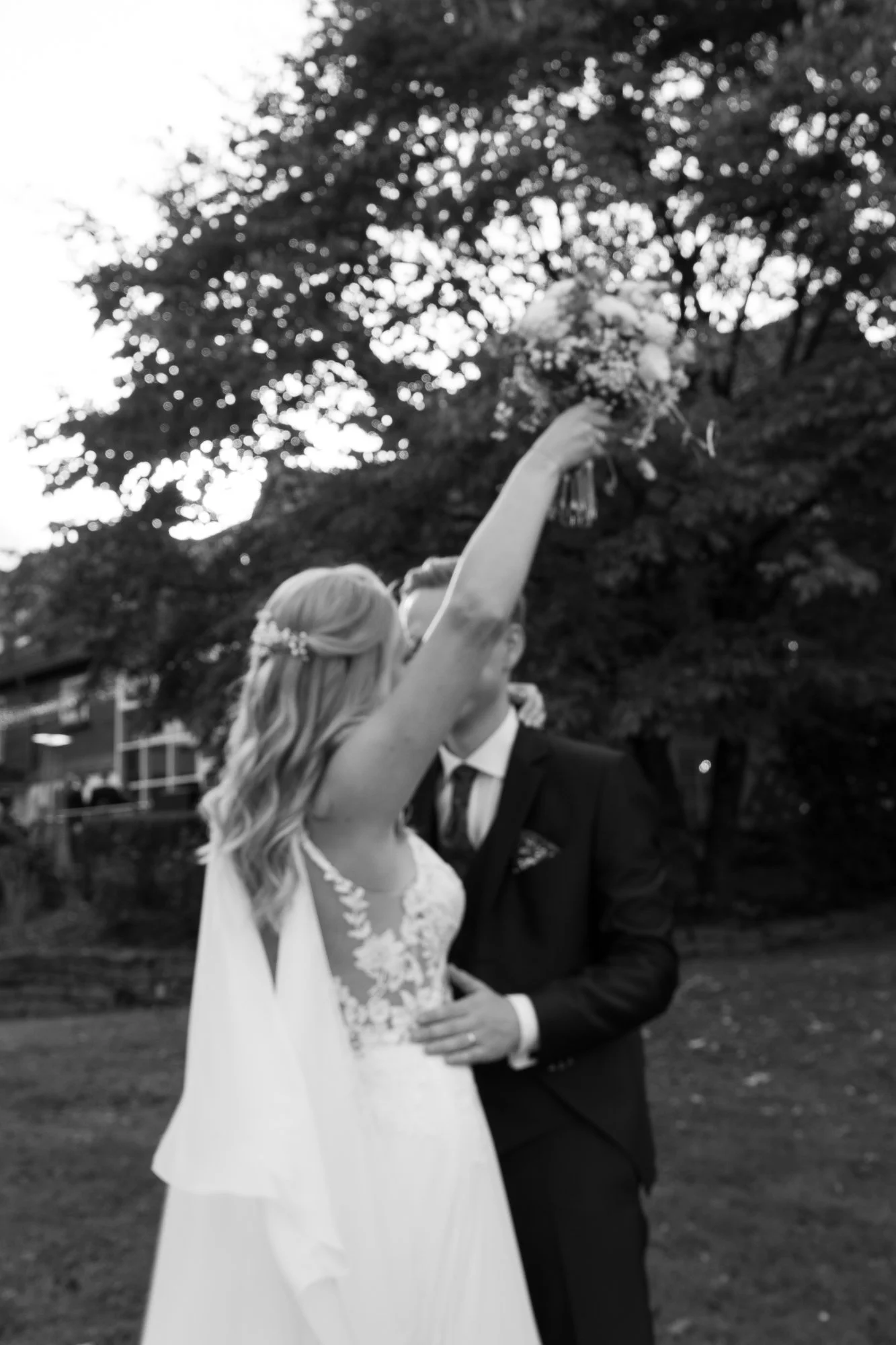 Braut hält einen Blumenstrauß hoch, während das Paar sich küsst, im Hintergrund Bäume, schwarz-weiß Aufnahme.