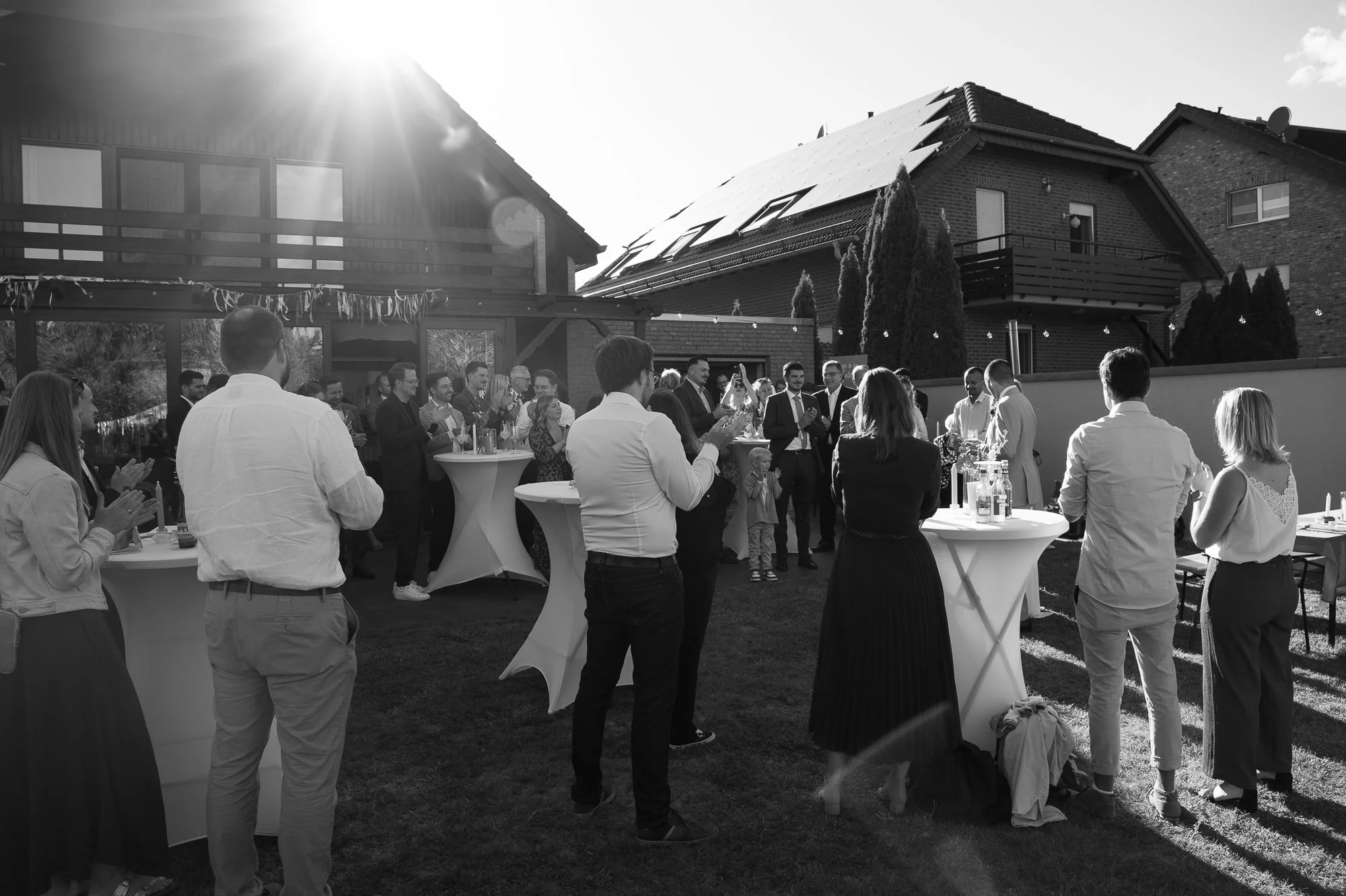 Eine Gruppe von Menschen beim Feiern im Garten eines Hauses bei Sonnenuntergang, mit Stehtischen und einem Buffet.