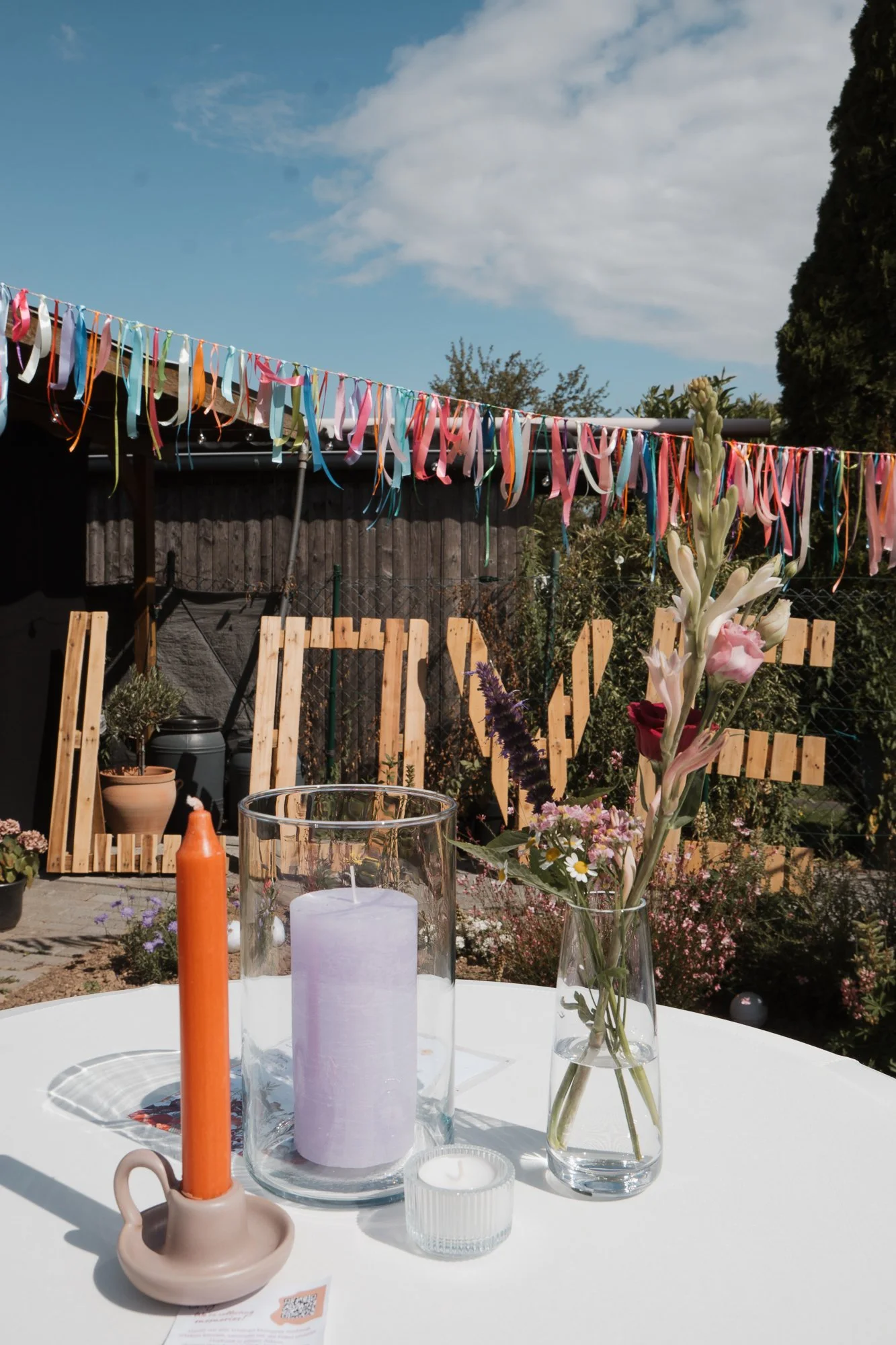 Blumenstrauß, Kerzen, Tischdeko bei einer Feier im Garten mit bunten Bändern im Hintergrund.