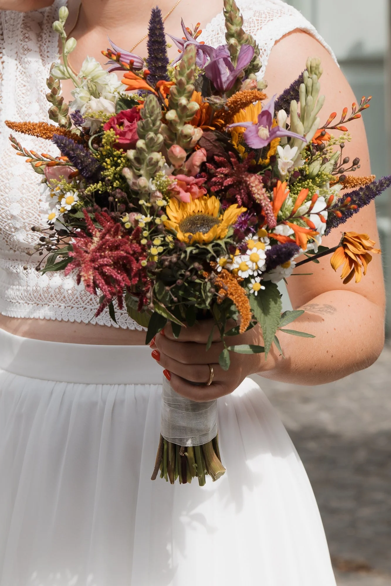 Frau in weißem Kleid hält einen bunten Blumenstrauß mit Sonnenblumen, Lavendel, Margeriten und anderen Blumen.