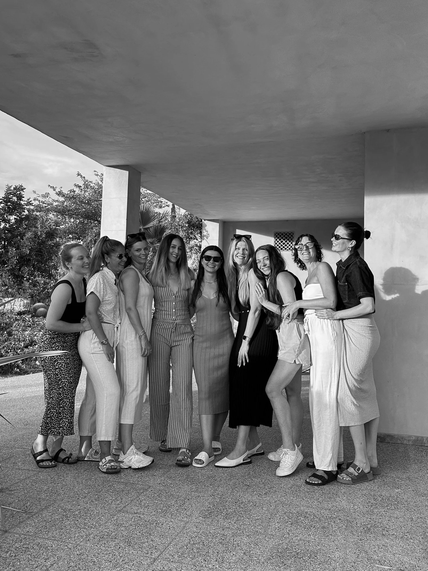 Eine Gruppe weiblicher Freunde posiert lachend unter einer modernen Architektur mit einem schattigen Bereich, im Freien, bei sonnigem Wetter.