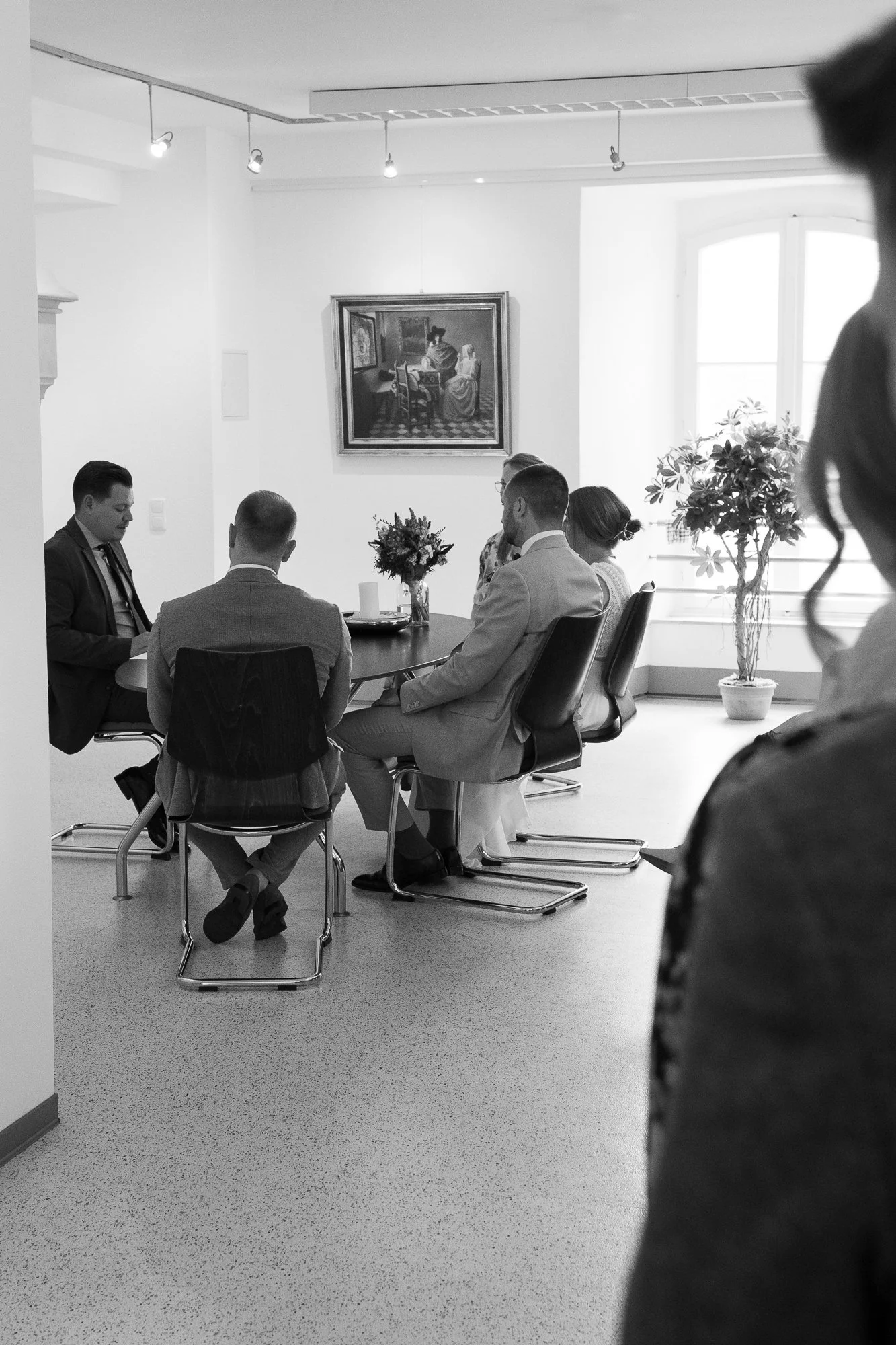 Fünf Menschen sitzen an einem runden Tisch in einem hellen Raum, vermutlich bei einer Zeremonie. Es gibt eine Vase mit Blumen auf dem Tisch und eine große Pflanze am Fenster. An der Wand hängt ein Gemälde.