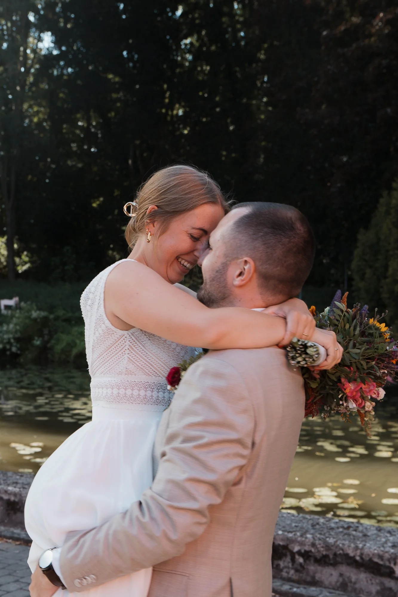 Ein lachendes Paar, das sich umarmt, bei einer Hochzeit im Freien mit Wasser im Hintergrund.