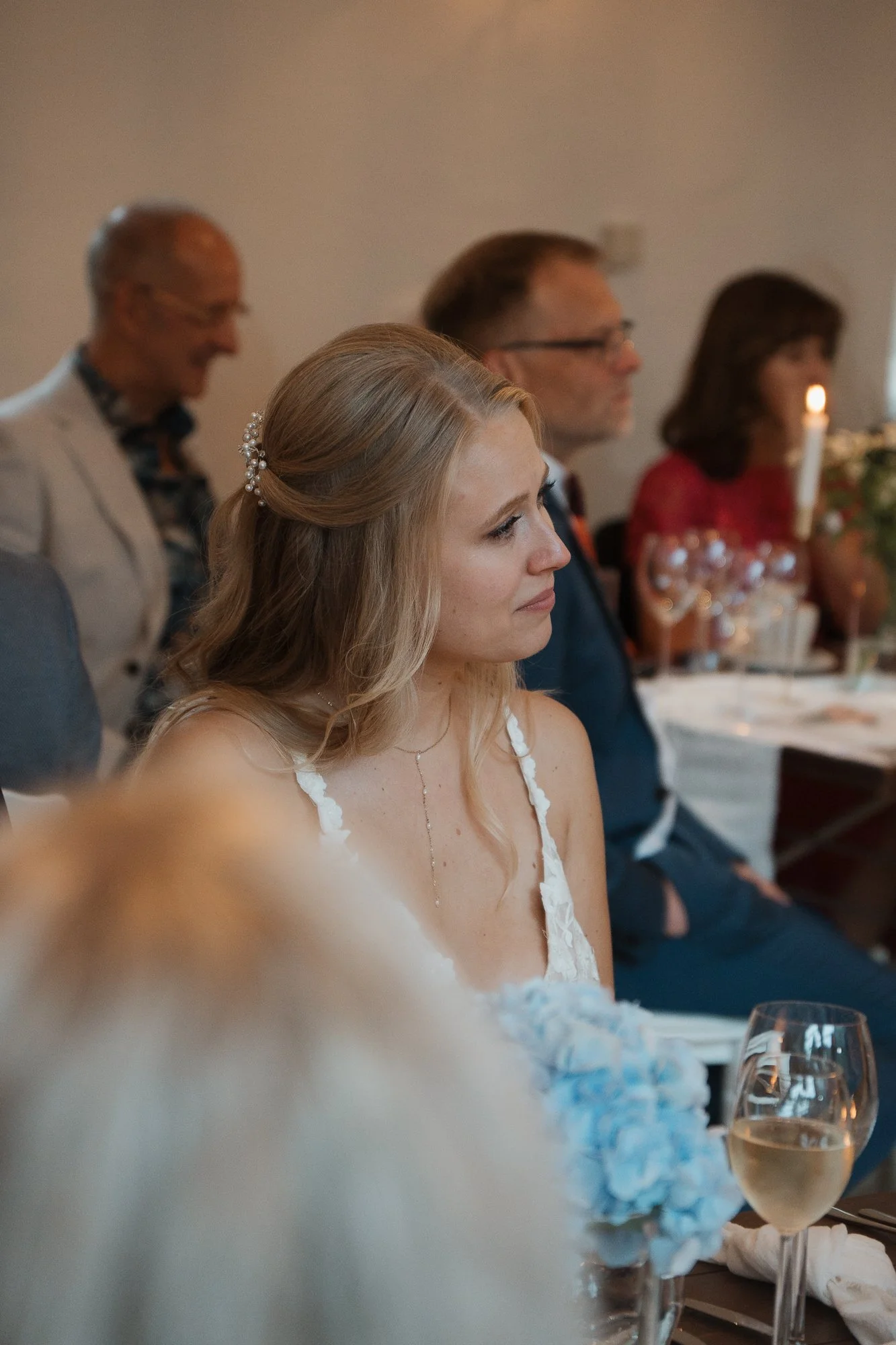 Junge Frau bei einer Hochzeit, sitzend am Tisch mit Blumen und einem Glas Wein, in einem festlichen Raum.