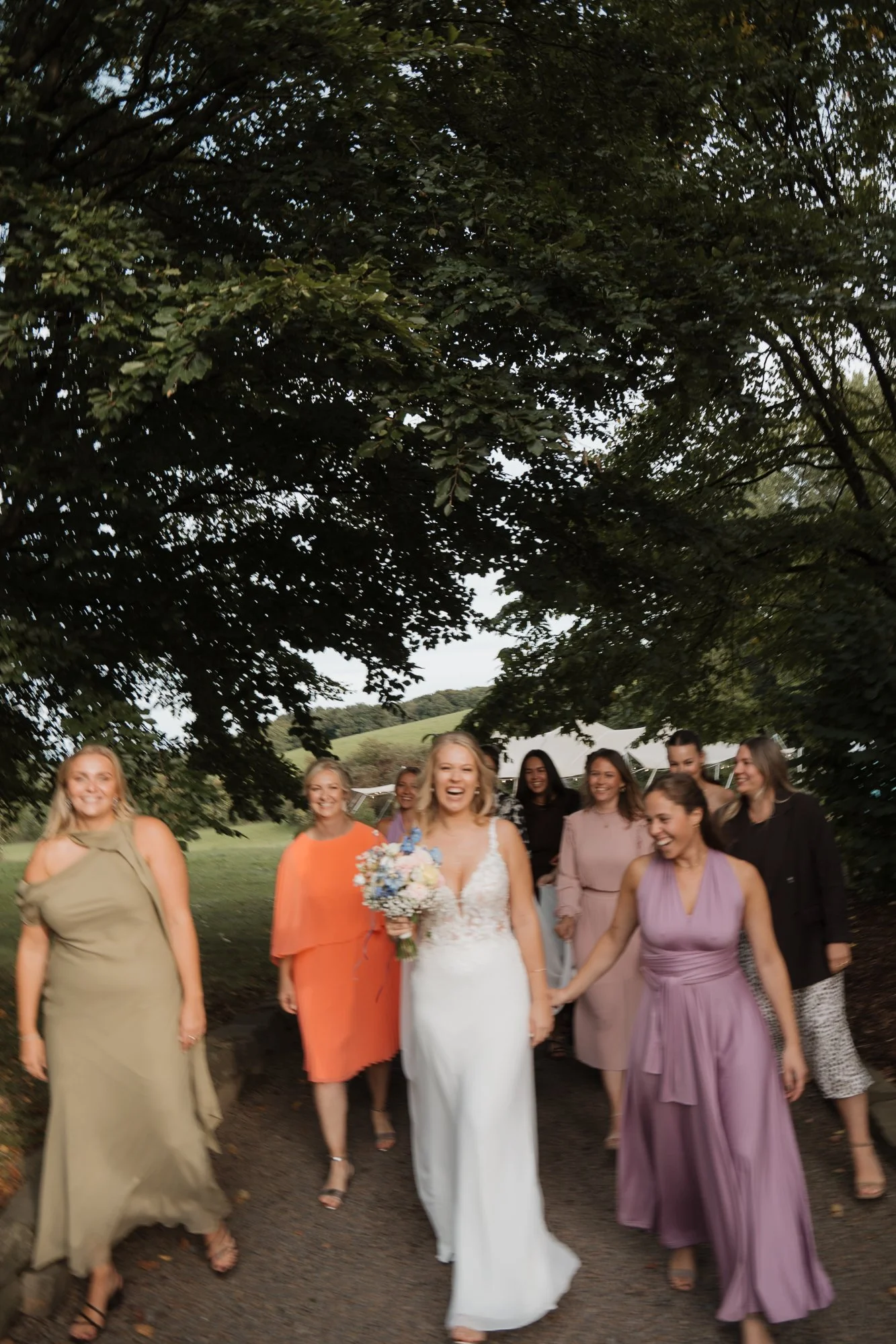 Eine Gruppe lachender Frauen bei einer Hochzeit, eine Frau in einem weißen Brautkleid mit einem Blumenstrauß in der Hand führt die Gruppe an, unter einem großen Baum im Freien.