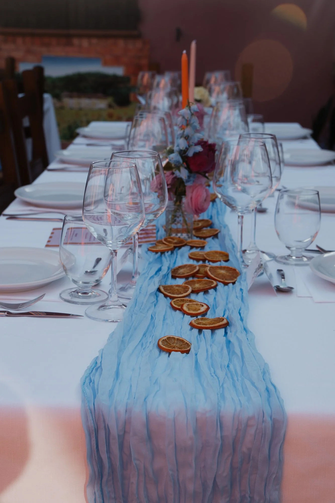 Ein festlich gedeckter Tisch mit weißen Tellern, Weingläsern, blauen Stoffservietten, einer Blütenmitte mit Kerzen und getrockneten Orangenscheiben als Tischdekoration.