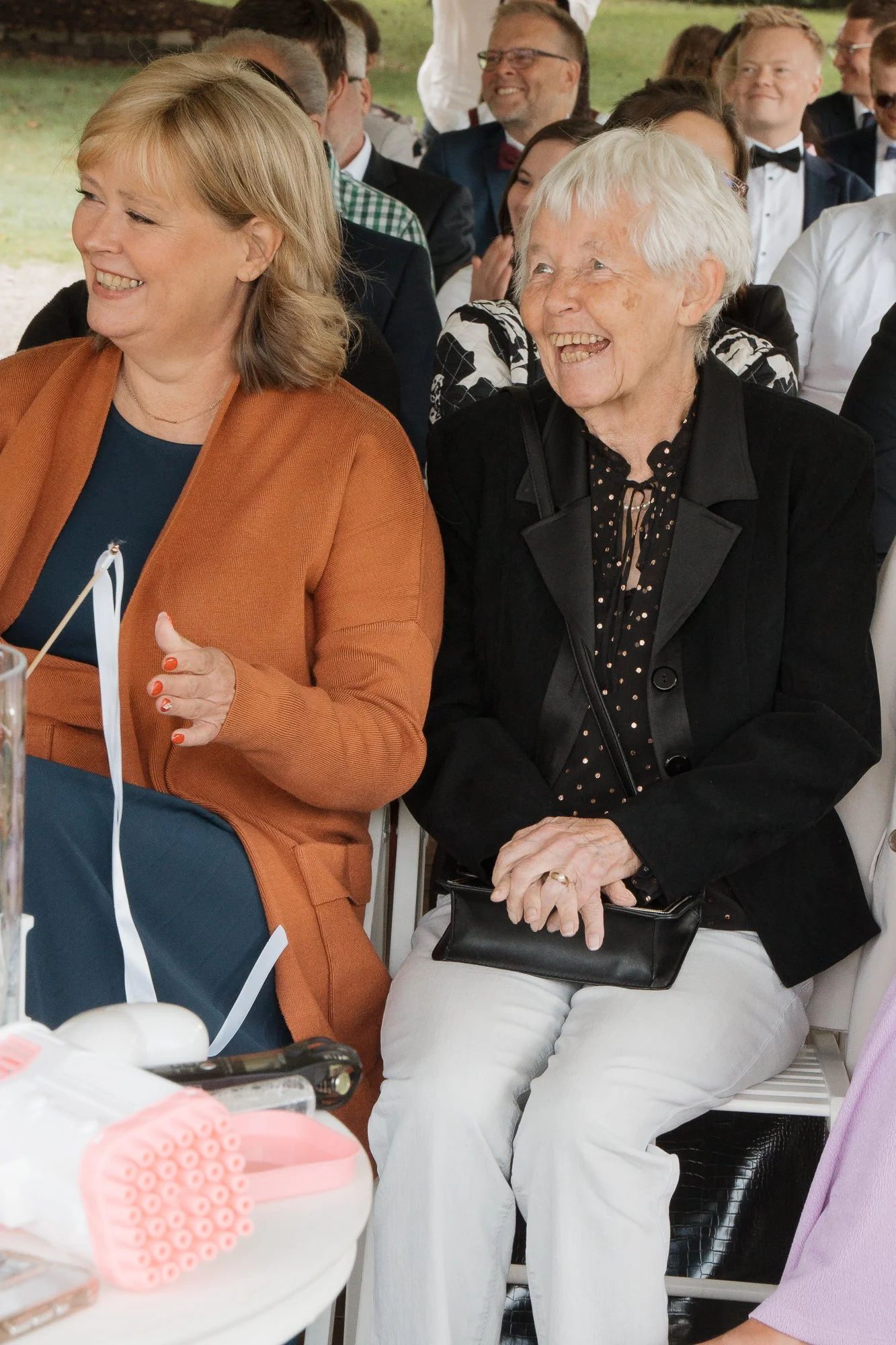 Menschen lachen und lachen bei einer Veranstaltung, im Vordergrund zwei Frauen, eine ältere mit weißen Haaren und dunkelblasem Anzug, die andere mit blonden Haaren und brauner Jacke