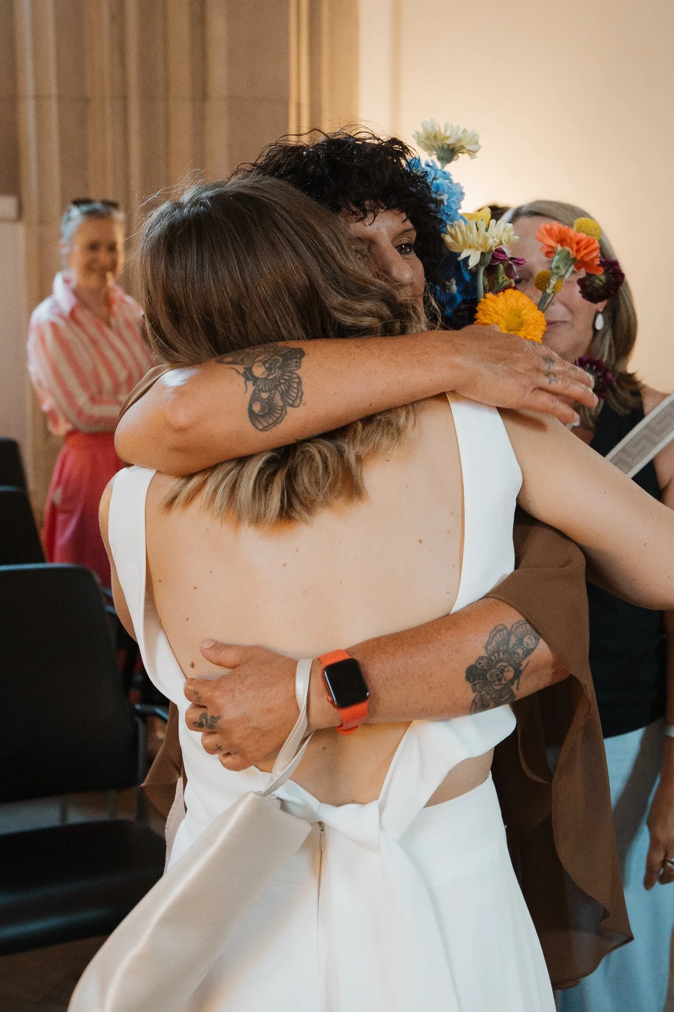 Zwei Frauen umarmen sich herzlich bei einer Feier, mit Blumen im Haar, im Hintergrund andere Gäste.