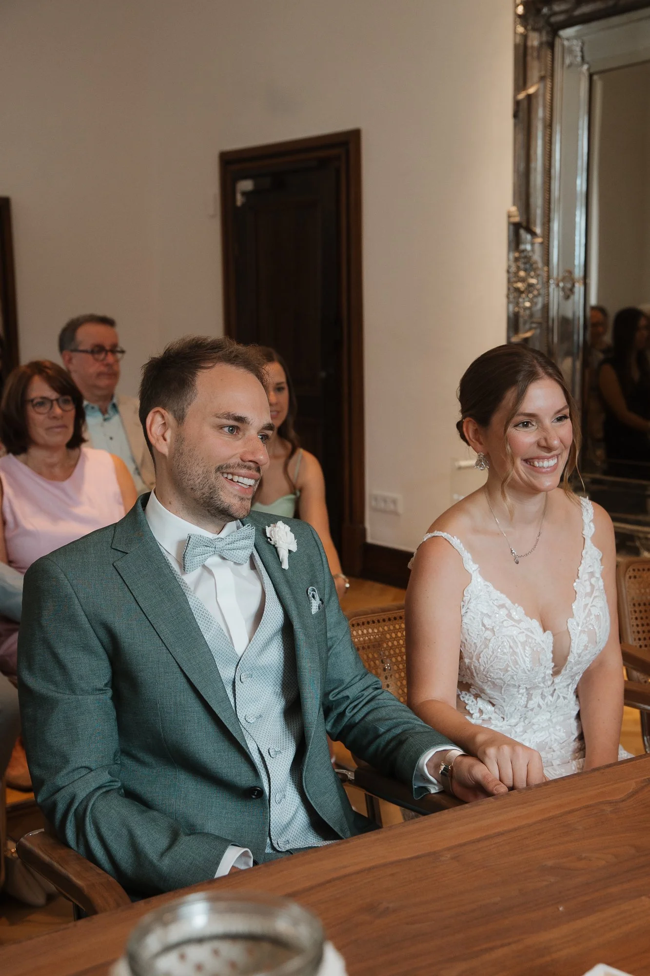 Braut und Bräutigam sitzen während ihrer Hochzeit in einem festlichen Raum, umgeben von Gästen.