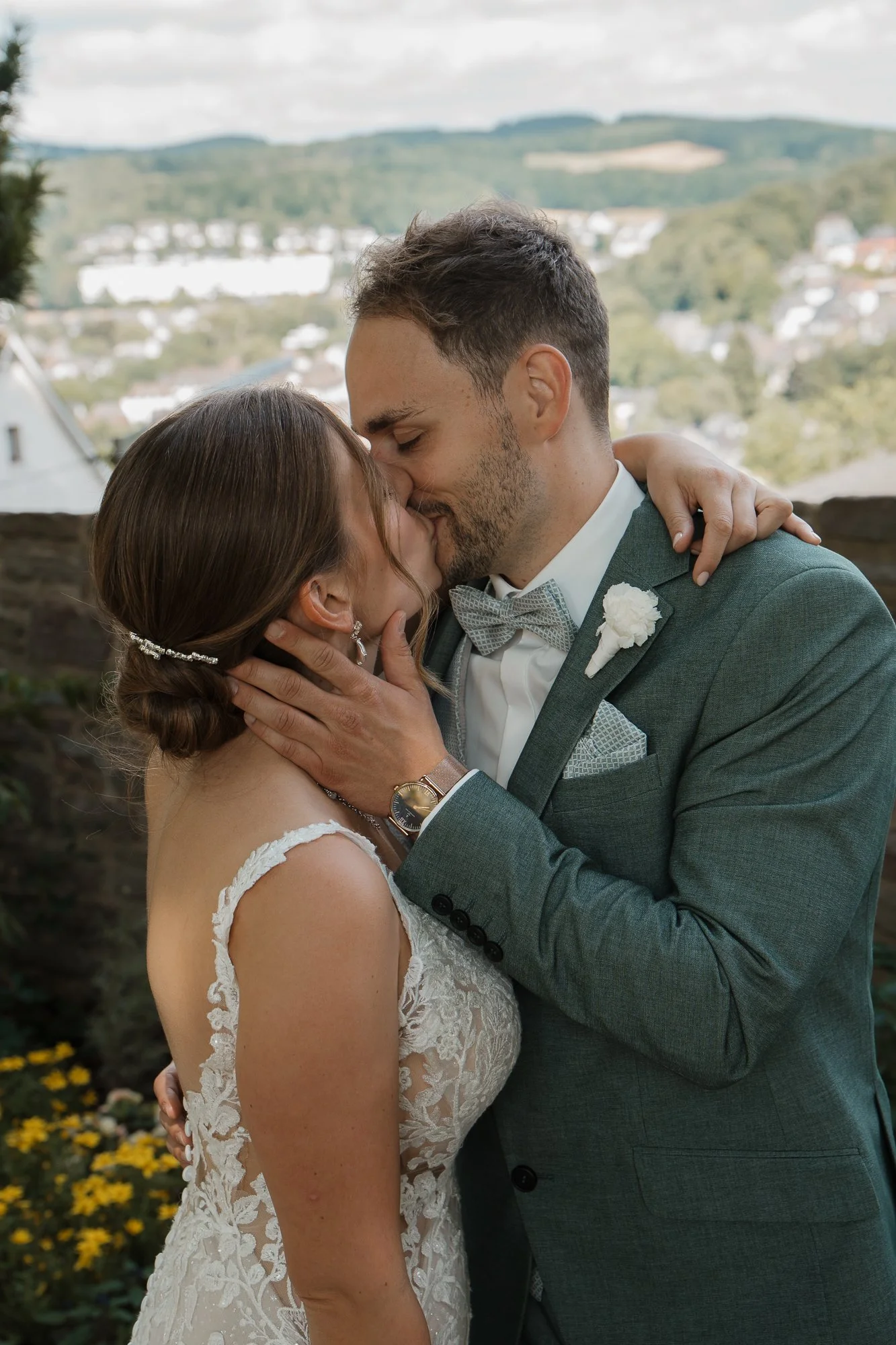 Ein Brautpaar küsst sich bei ihrer Hochzeit im Freien, im Hintergrund sieht man ein ländliches Gebiet mit Häusern und Hügel.