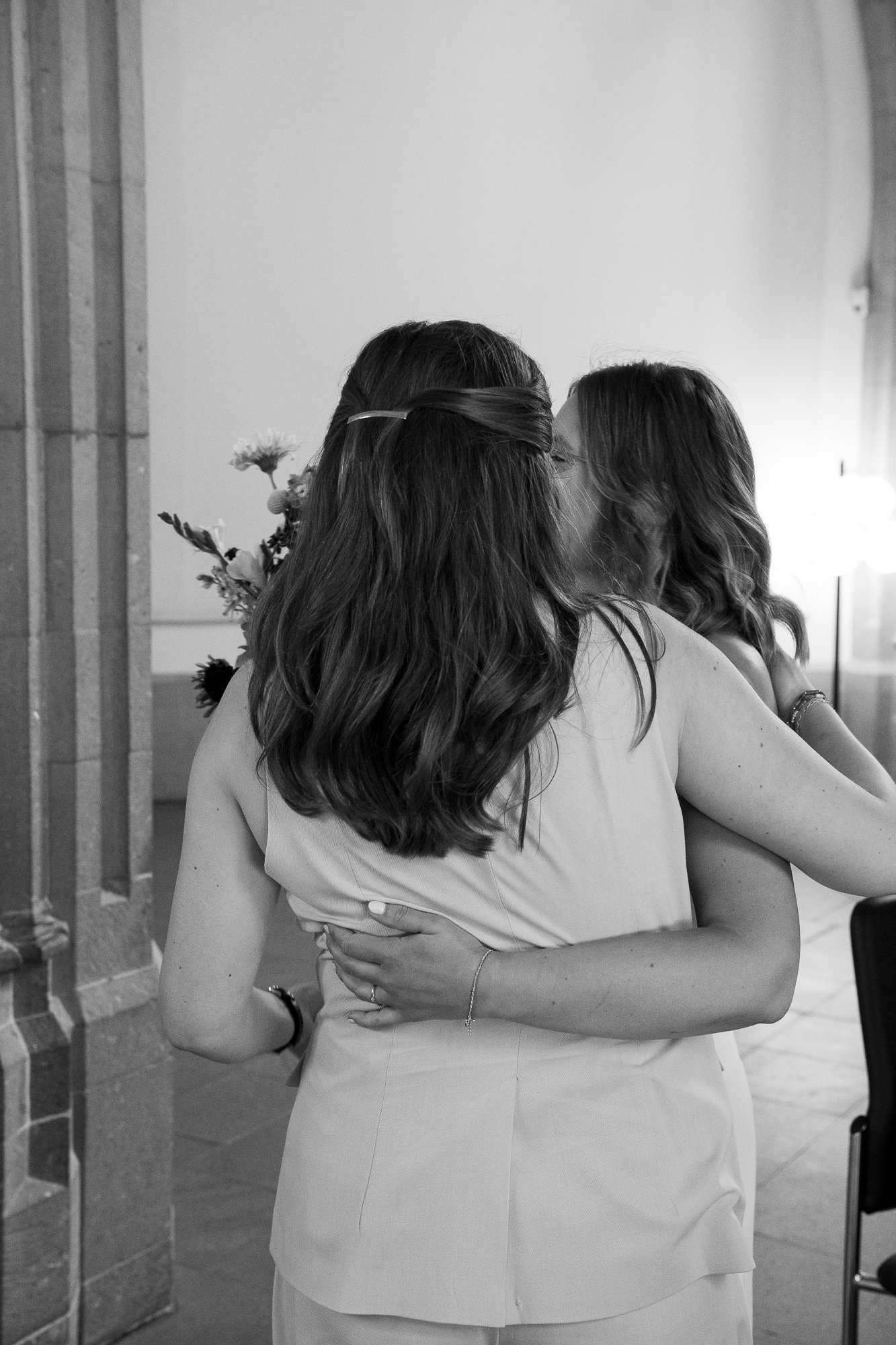 Zwei Frauen umarmen sich bei einer Zeremonie, eine Frau küsst die andere auf die Wange, im Hintergrund eine weitere Person mit Blumen.