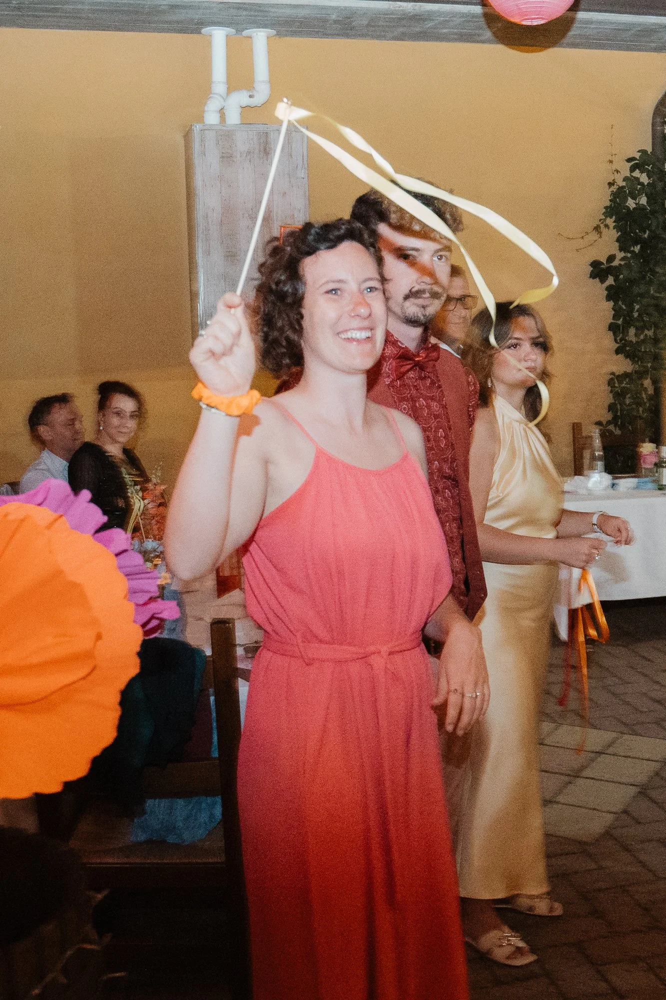 Mehrere Menschen feiern bei einer festlichen Veranstaltung. Eine Frau in einem roten Kleid hält eine goldene Spirale und lächelt in die Kamera. Hinter ihr stehen weitere Frauen in eleganten Kleidern und Menschen, die am Tisch sitzen.