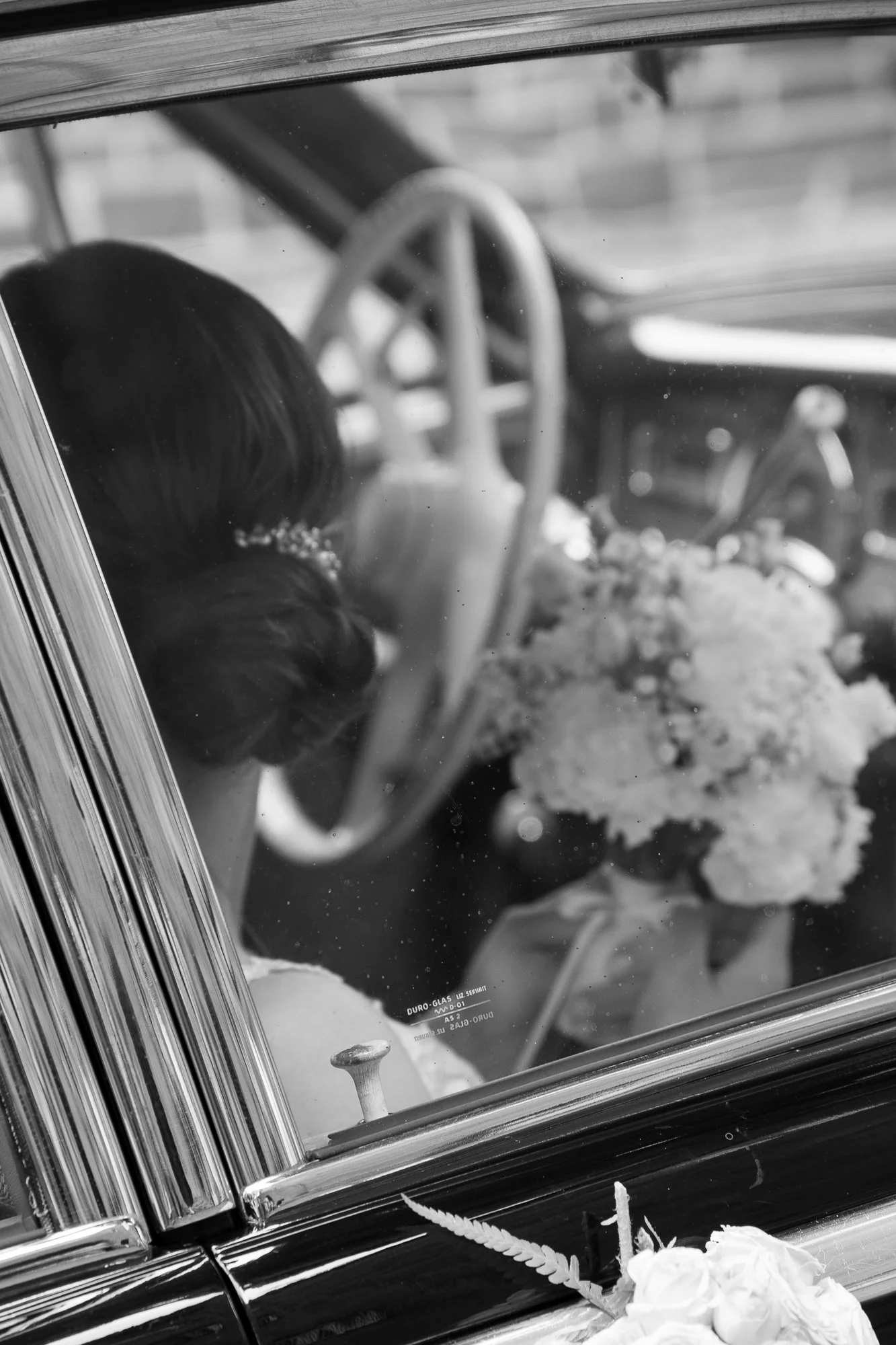 Schwarz-Weiß-Foto eines Brautpaares im Auto, mit Brautstrauß und Blumen vor dem Fahrzeug