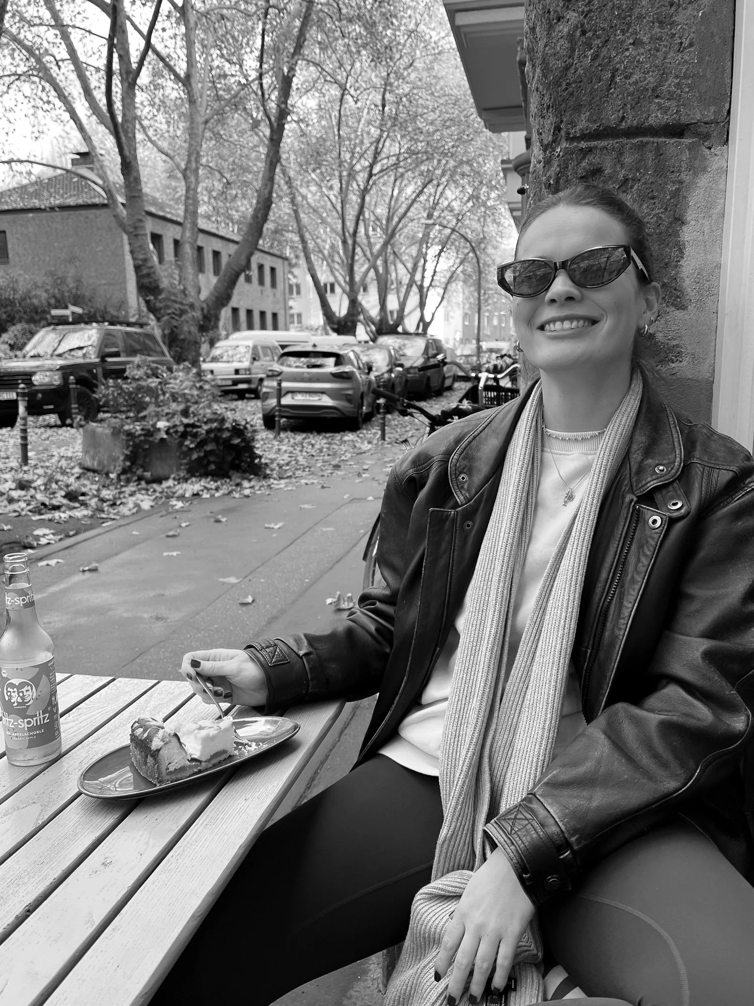 Eine Frau sitzt im Freien an einem Tisch, trägt Sonnenbrille und Lederjacke, hat eine Torte und ein Getränk vor sich, lächelt.