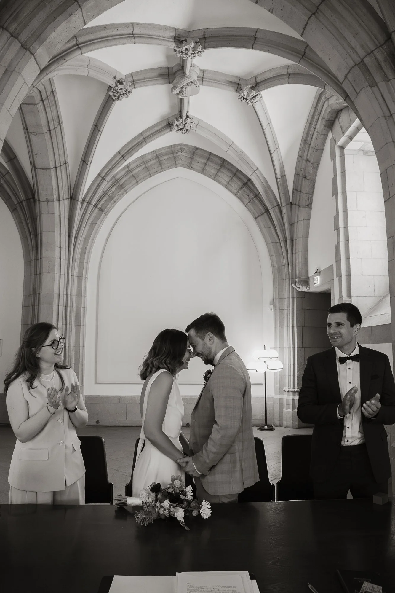 Ein Brautpaar hält sich bei einer Hochzeit im Sakralbau die Hände und schaut in die Augen, während ihre Freunde applaudieren.