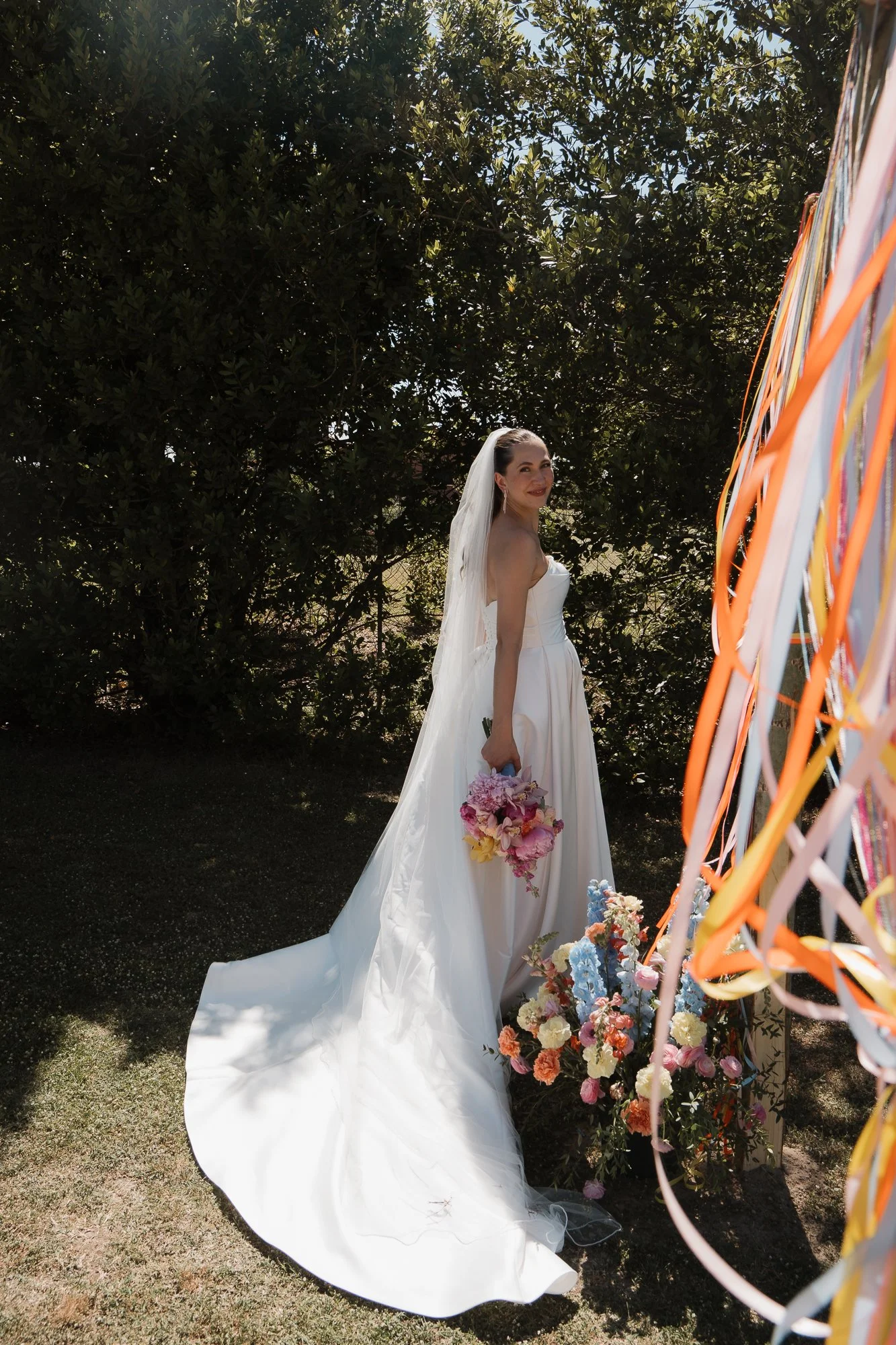 Eine Braut in weißem Brautkleid mit Schleier, die einen Blumenstrauß hält, steht im Freien vor Bäumen. Es ist sonnig, und sie lächelt in die Kamera, während sie neben farbigen Blumen und einem dekorativen Element mit bunten Bändern steht.