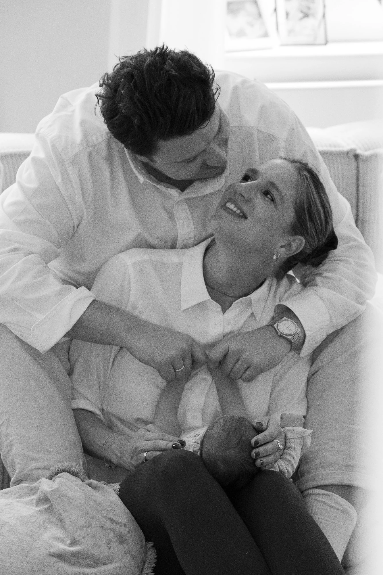 Ein Paar hält ein neugeborenes Baby im Arm und lächelt sich liebevoll an, während sie in einem Raum sitzen.