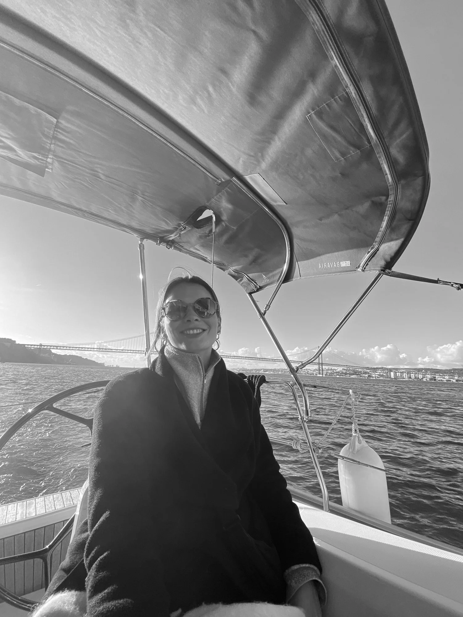 Eine Frau mit Sonnenbrille und dunklem Mantel sitzt auf einem Boot auf dem Wasser, im Hintergrund eine Brücke und ein Himmel mit Wolken.