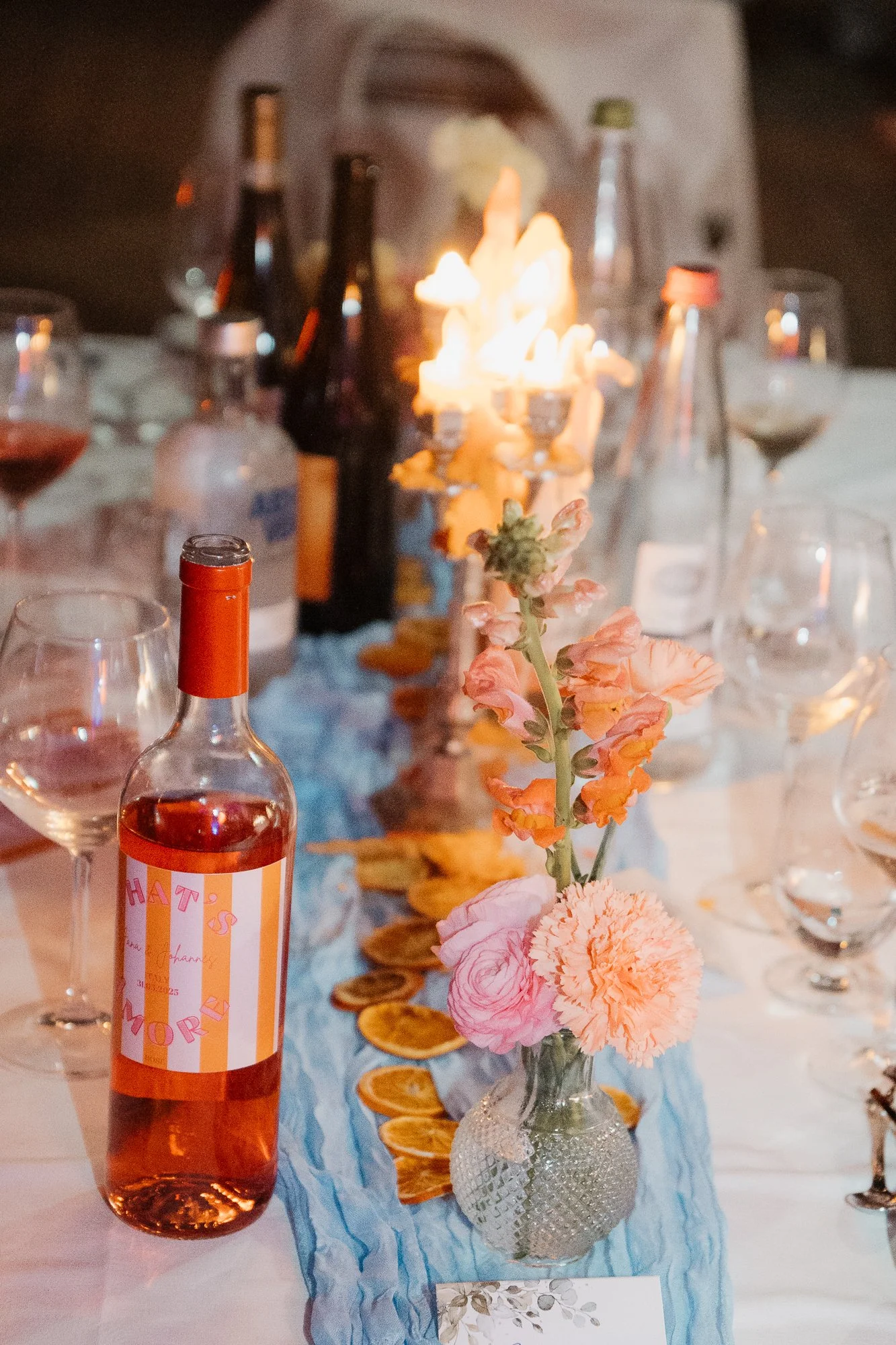 Tischdekoration mit pinken Blumen, einer Flasche Roséwein, Gläsern, einem Kerzenständer und getrockneten Orangenscheiben bei einem festlichen Abendessen.
