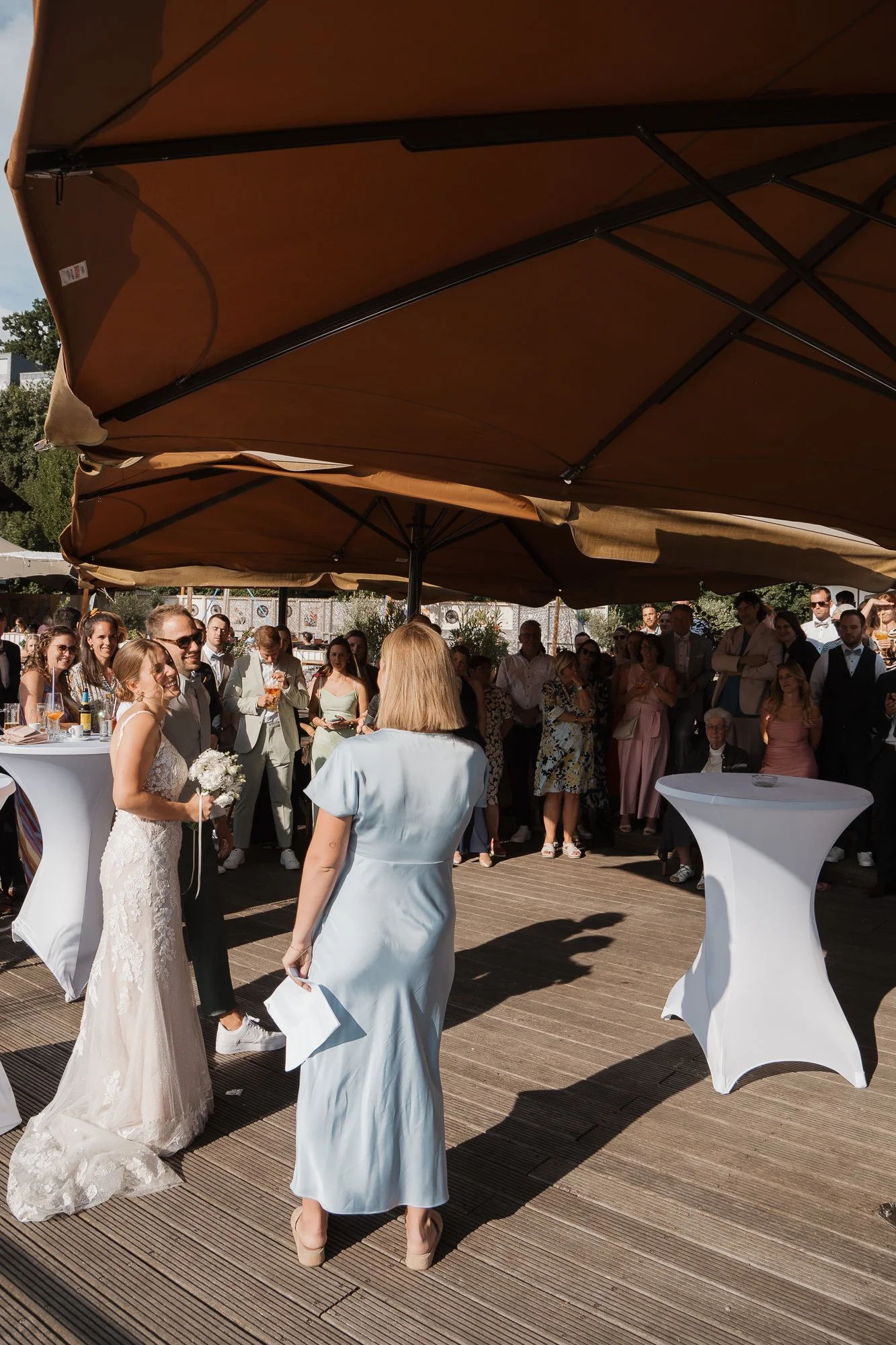 Hochzeit im Freien mit Braut und einem Gast, die sich unter großen Sonnenschirmen befinden, umgeben von einer Gruppe von Gästen.