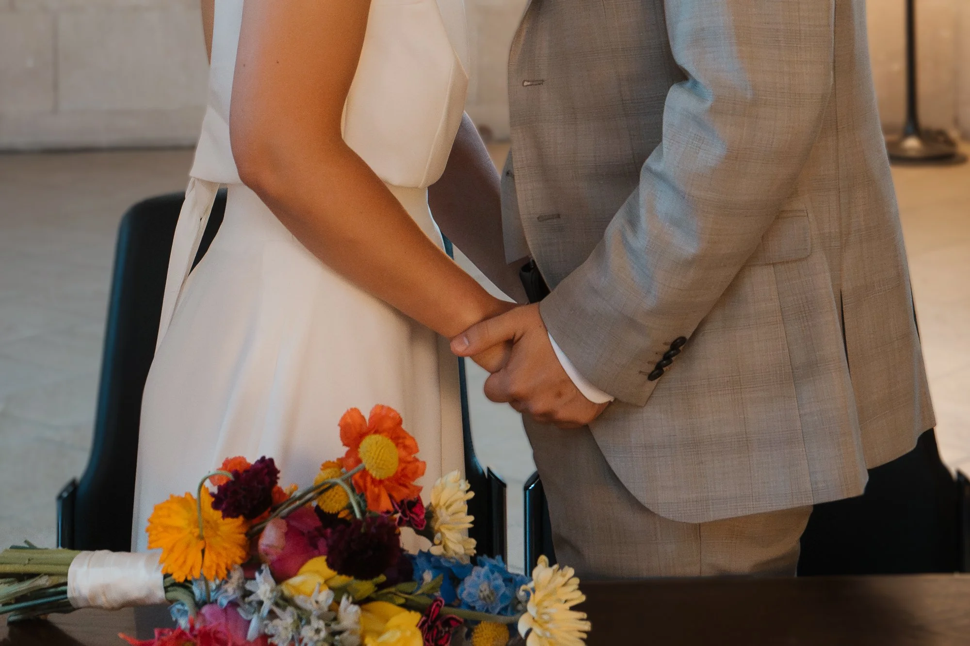 Hochzeitspaar hält Händchen bei Standesamt, Blumenstrauß liegt auf Tisch.