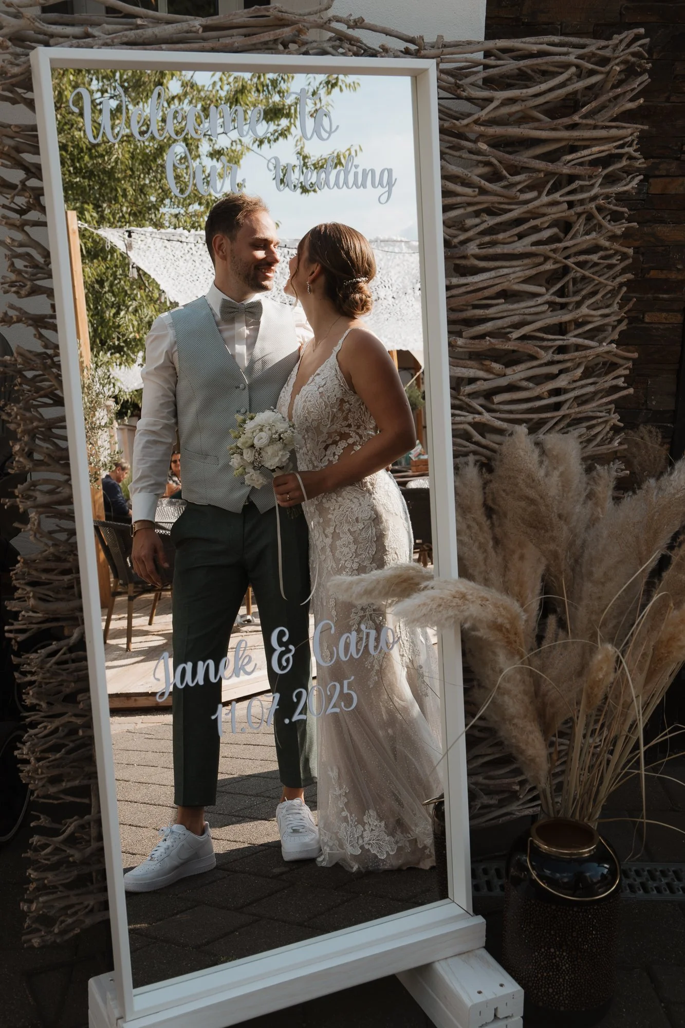 Ein Spiegel mit der Aufschrift 'Willkommen zu unserer Hochzeit' spiegelt ein Brautpaar, das sich liebevoll anschaut, vor einem ländlichen Hintergrund mit Bäumen und Dekorationen. Das Brautpaar besteht aus einem Mann in Anzug und einer Frau in einem e