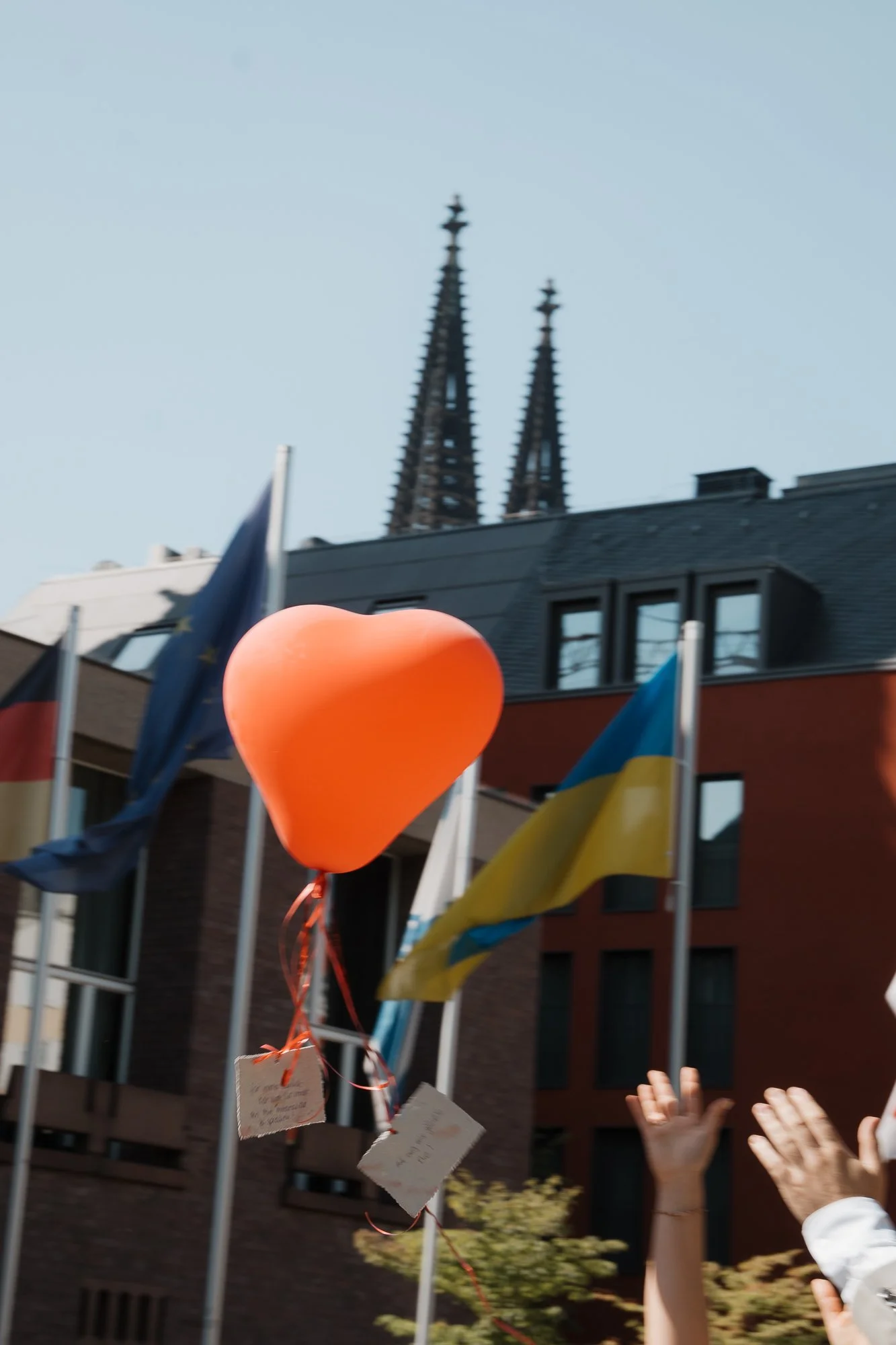 Ein orangefarbener Luftballon in Herzform, der an einer Schnur mit kleinen Zetteln befestigt ist, im Vordergrund, umgeben von Menschen mit erhobenen Händen, dahinter Flaggen einer EU, eines Landes und einer Ukraine, und im Hintergrund die Türme der S