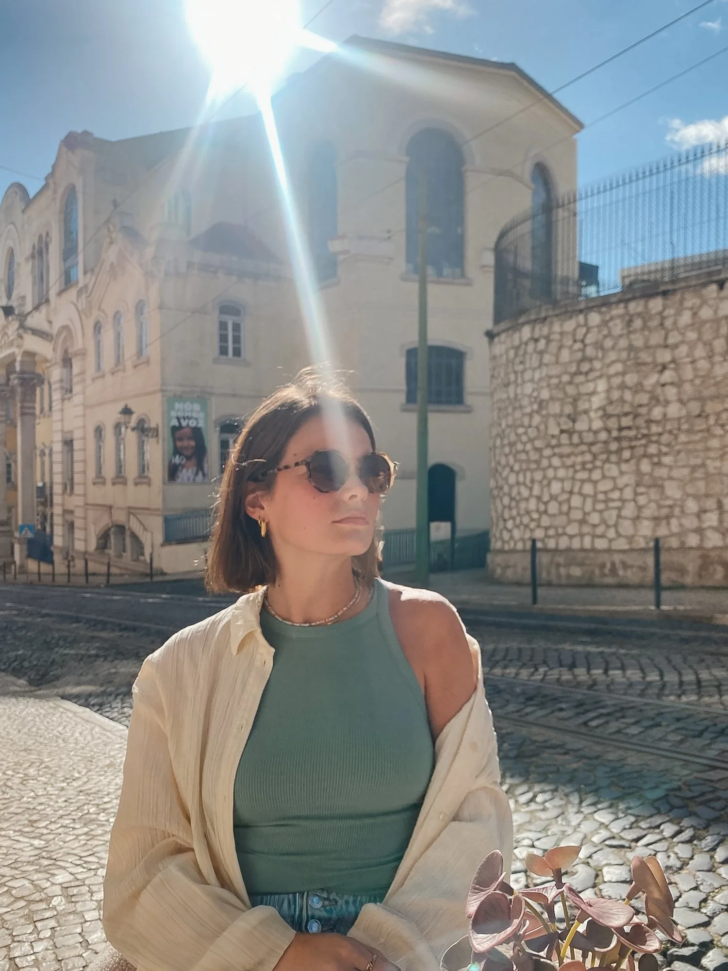 Junge Frau mit kurzen braunen Haaren trägt runde Sonnenbrille, eine türkisfarbene Tanktop und eine offene cremefarbene Jacke, sitzt auf einem Kopfsteinpflaster in der Sonne vor alten Gebäuden in einer Stadt, während die Sonne hell am Himmel scheint.