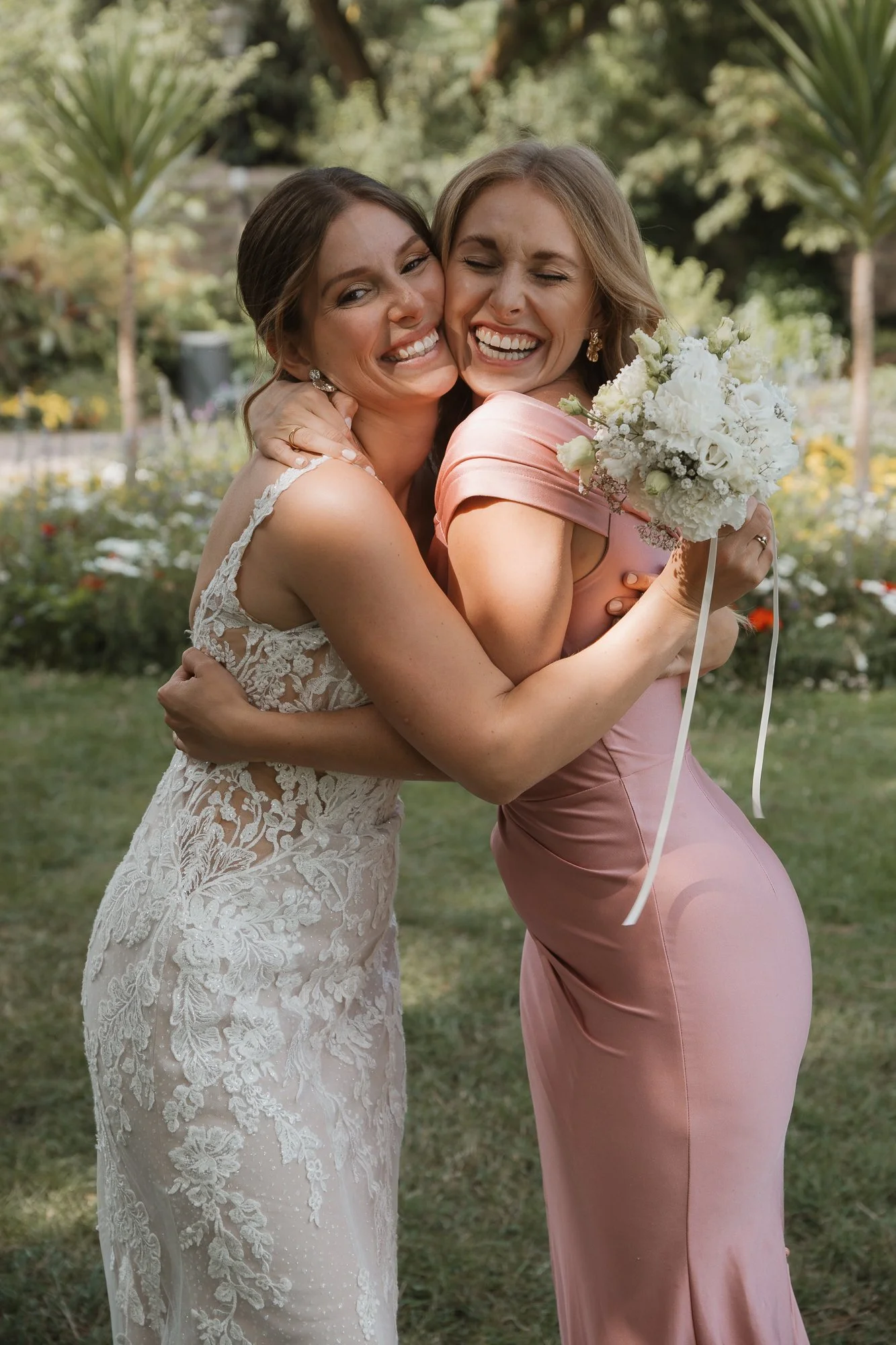 Zwei Frauen im Freien, die sich umarmen, eine trägt ein weißes Hochzeitskleid, die andere ein rosa Kleid und hält einen Brautstrauß.
