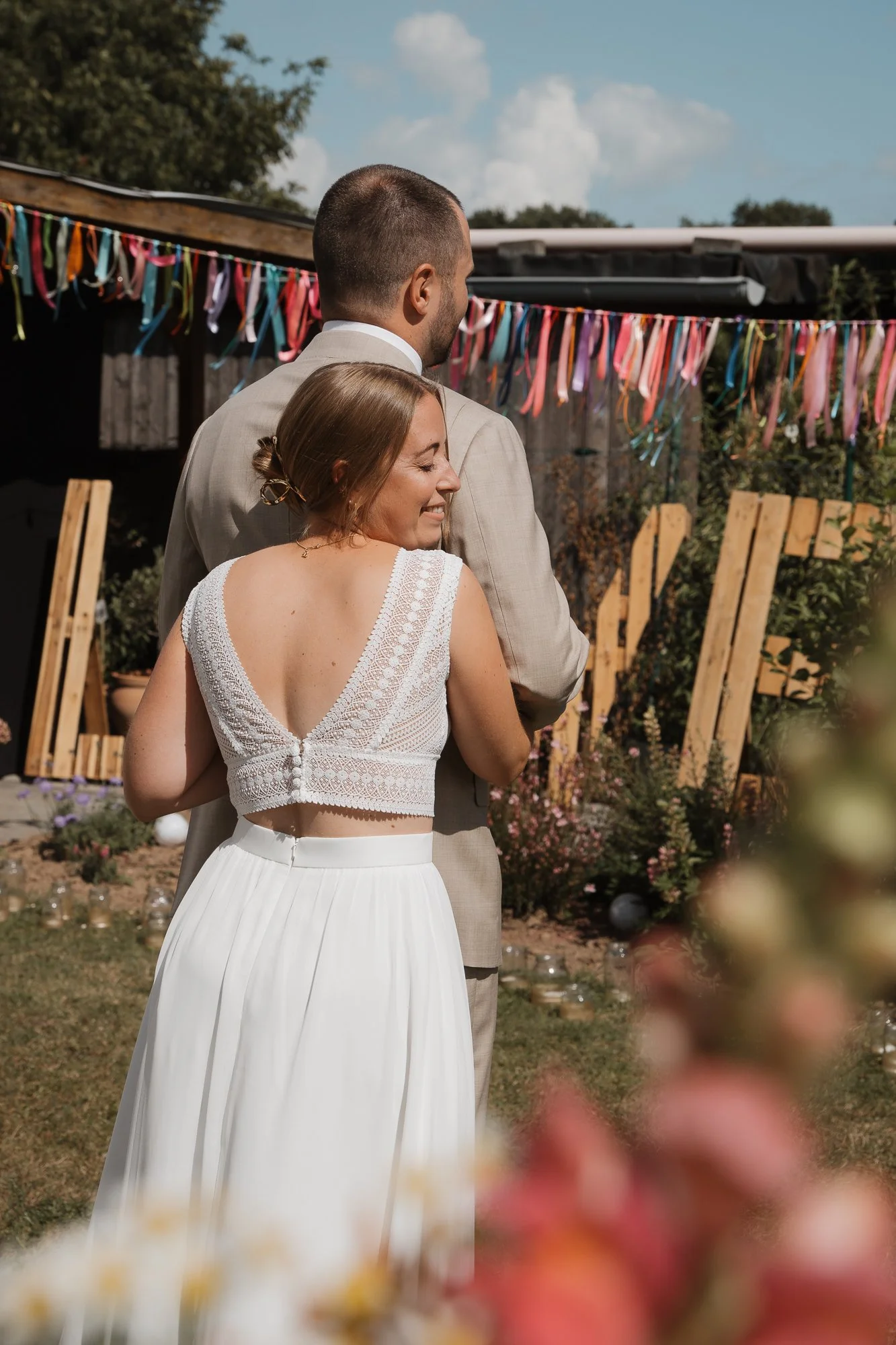 Ein Paar bei einer Hochzeit im Garten, die Frau trägt ein weißes Kleid mit Spitze, der Mann einen beigen Anzug, bunte Bänder im Hintergrund