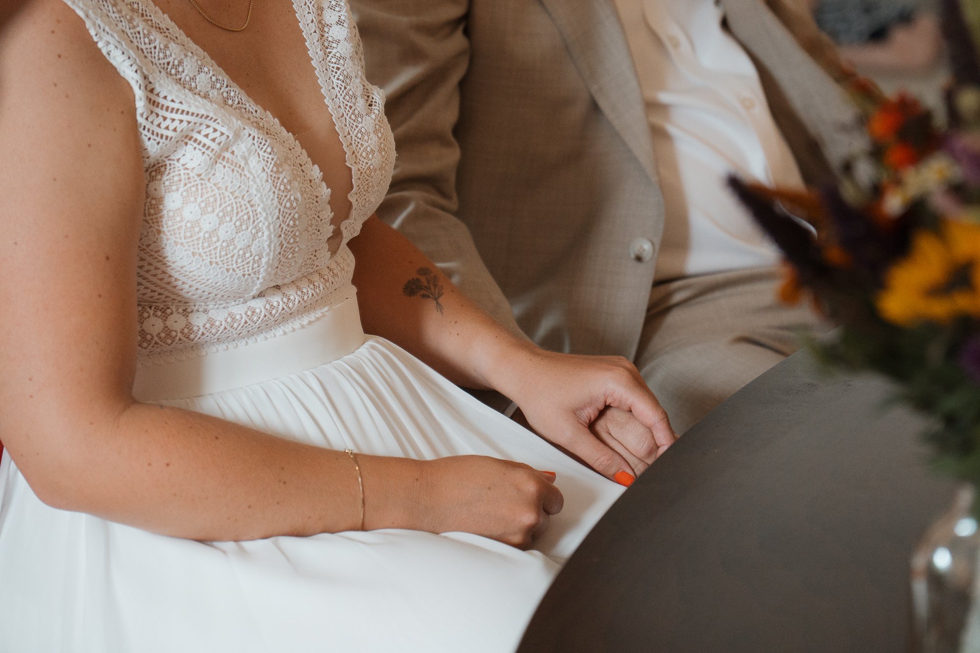 Nahaufnahme einer Braut und eines Bräutigams bei einer Hochzeit, die auf einem grauen Tisch sitzen. Die Braut trägt ein weißes Spitzenkleid, der Bräutigam einen hellgrauen Anzug. Eine Blumenarrangement ist im Vordergrund sichtbar.
