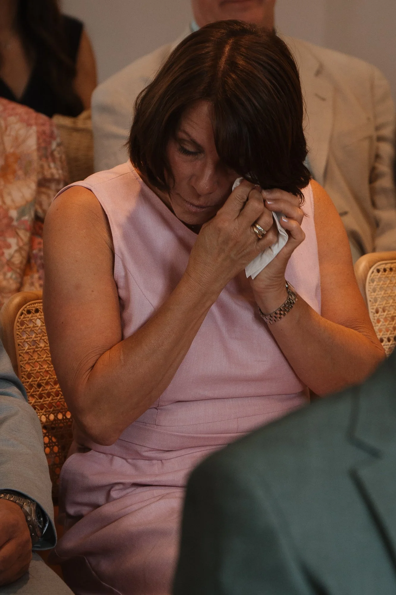 Frau in pinkem Kleid sitzt und weint, hält ein Taschentuch vor das Gesicht, in einem Raum mit anderen Personen.