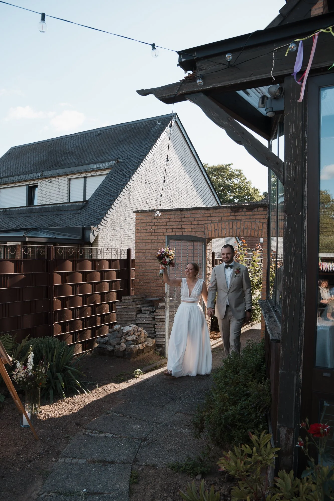 Ein Brautpaar geht Hand in Hand durch den Garten an einem Tag mit blauem Himmel. Die Braut trägt ein weißes Hochzeitskleid und hält einen Blumenstrauß hoch, während der Bräutigam einen hellgrauen Anzug mit schwarzer Krawatte trägt.