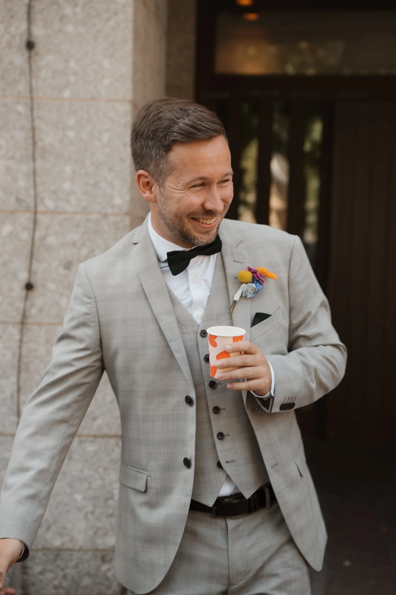 Ein Mann in einem hellgrauen Anzug, weißen Hemd, schwarzer Fliege und mit einer bunten Blume im Revers steht vor einer Steinwand und hält einen Pappbecher, während er lächelt.