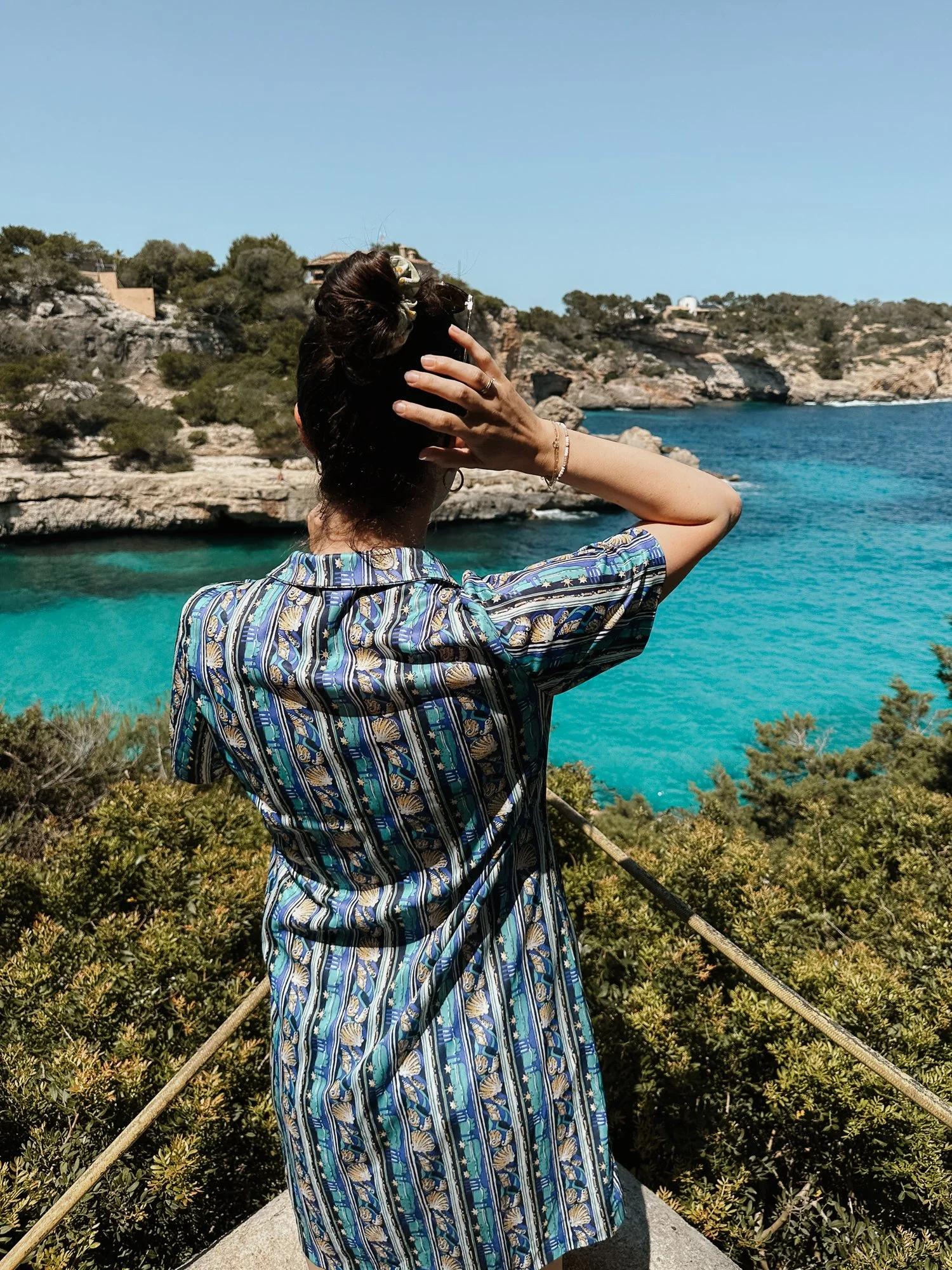 Frau mit geflecktem blauen Kleid, die auf einem Aussichtspunkt mit Blick auf das Blau des Meeres steht, die Haare zurückgebunden, während sie den Blick genießt.