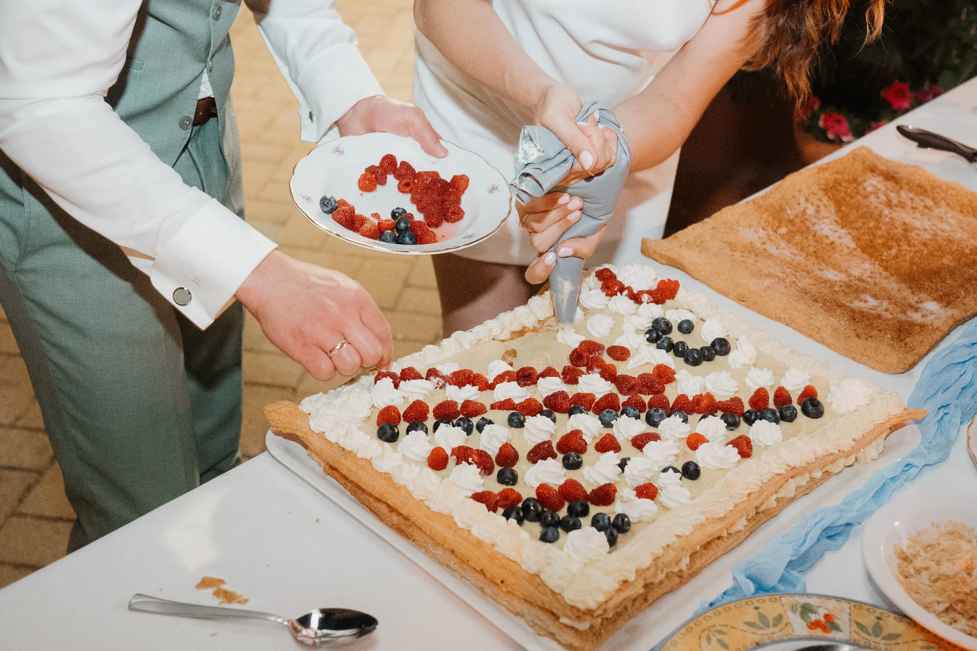 Menschen dekorieren eine große rechteckige Torte mit Sahne und Beeren auf einem festlich gedeckten Tisch.