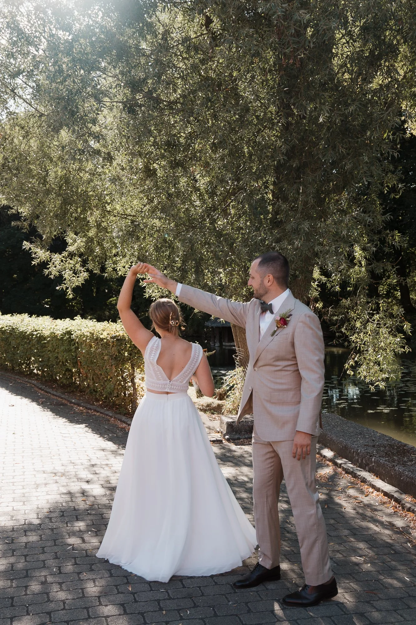 Ein Brautpaar tanzt im Freien bei Sonnenlicht, umgeben von Bäumen und Wasser. Der Bräutigam trägt einen hellen Anzug mit Fliege, die Braut in einem weißen Kleid mit tiefem Rücken.