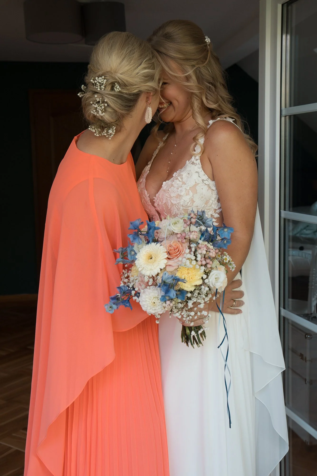 Zwei Frauen, vermutlich eine Braut und ihre Mutter, stehen sich nahe und lächeln sich an, während die Braut ein Blumenbouquet hält. Die Mutter trägt ein orangefarbenes Kleid, die Braut ein weißes Hochzeitskleid mit Spitzen-Details.