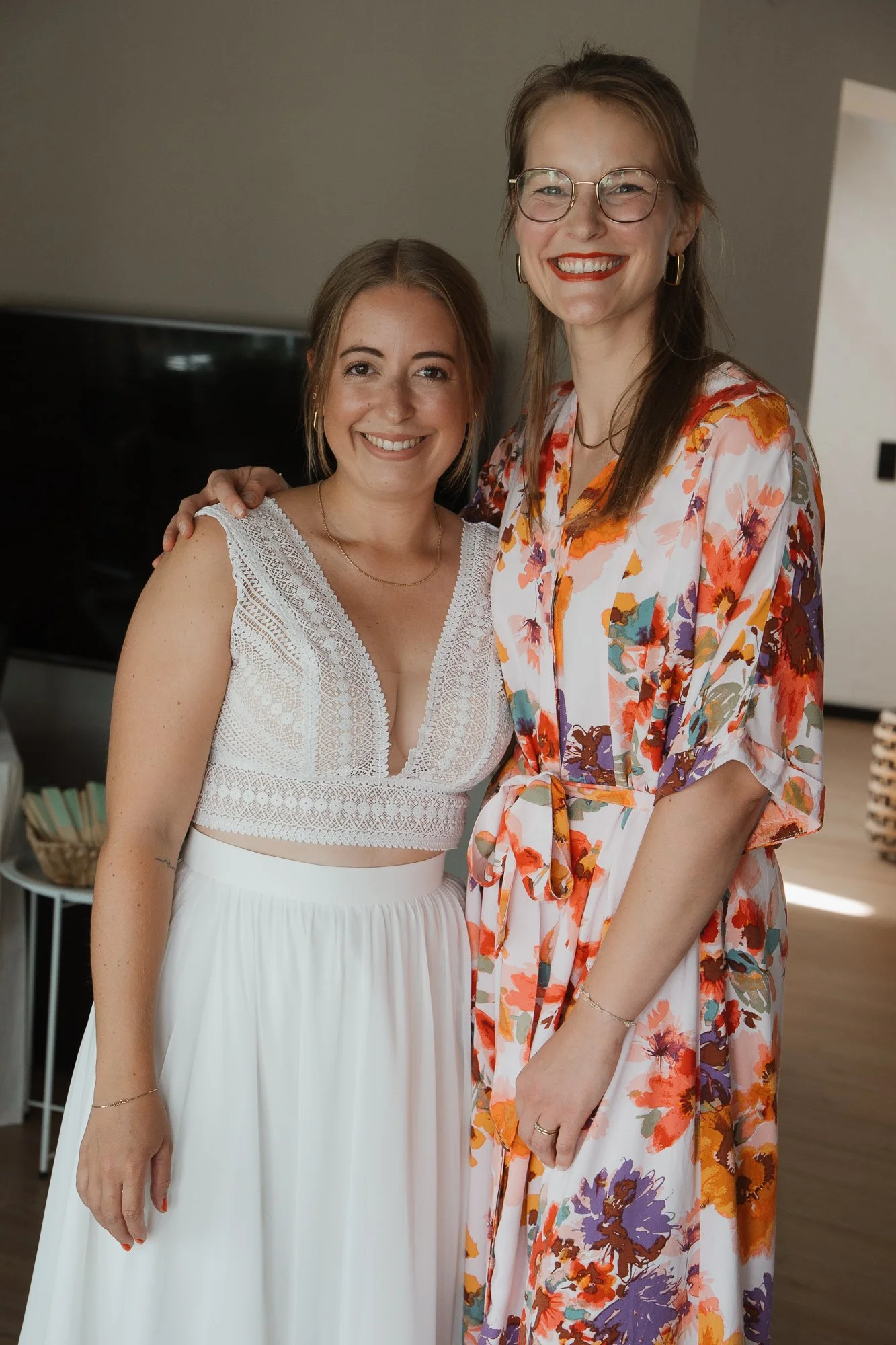 Zwei lachende Frauen stehen zusammen in einem Zimmer, eine trägt ein weißes kleid mit spitzen Details, die andere ein buntes sommerliches Kleid mit Blumenmuster.