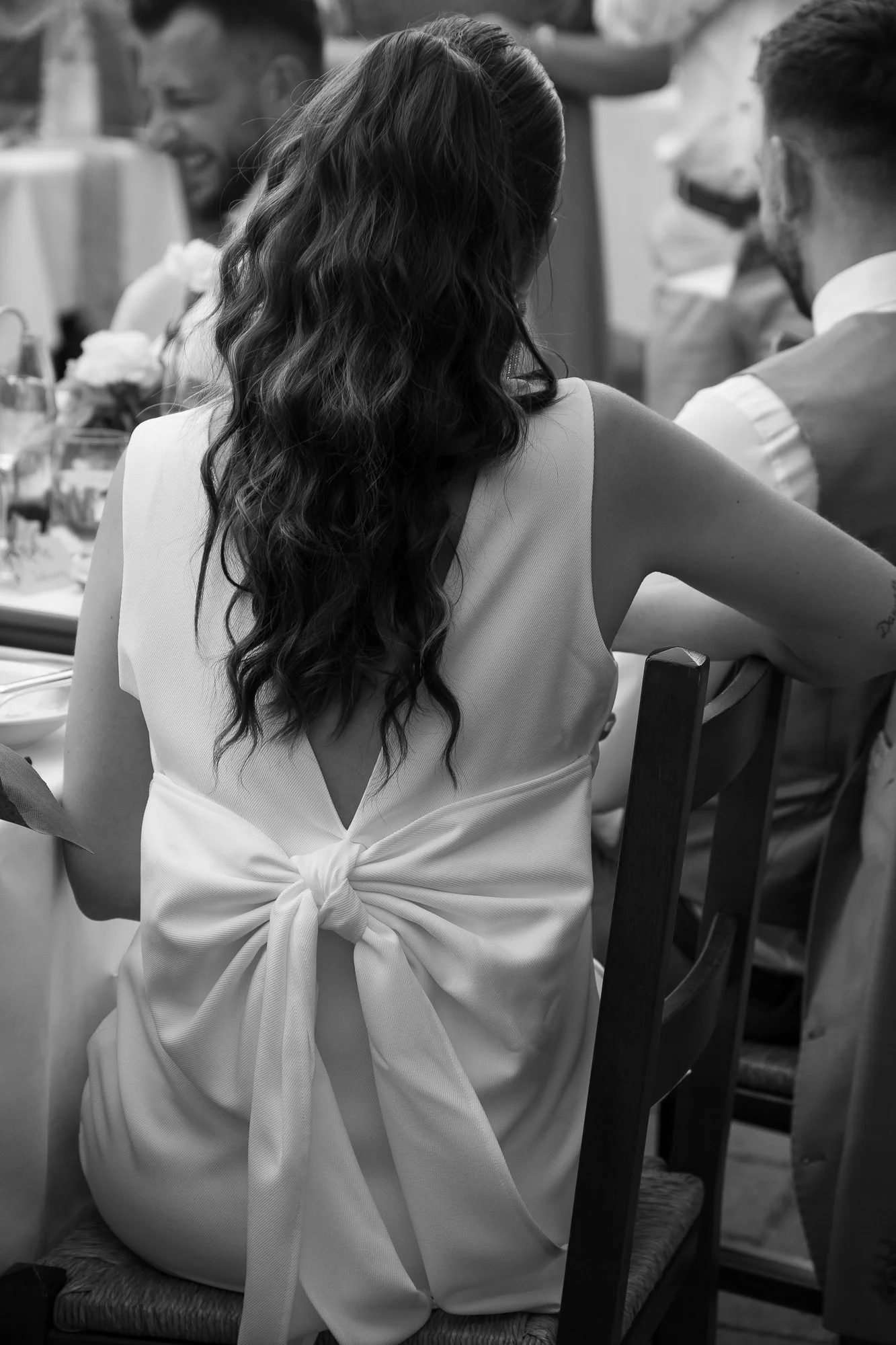 Frau mit langen, lockigen Haaren trägt ein weißes Kleid mit offener Rückenpartie und Schleife am Rücken, sitzt an einem Tisch bei einer gesellschaftlichen Veranstaltung.